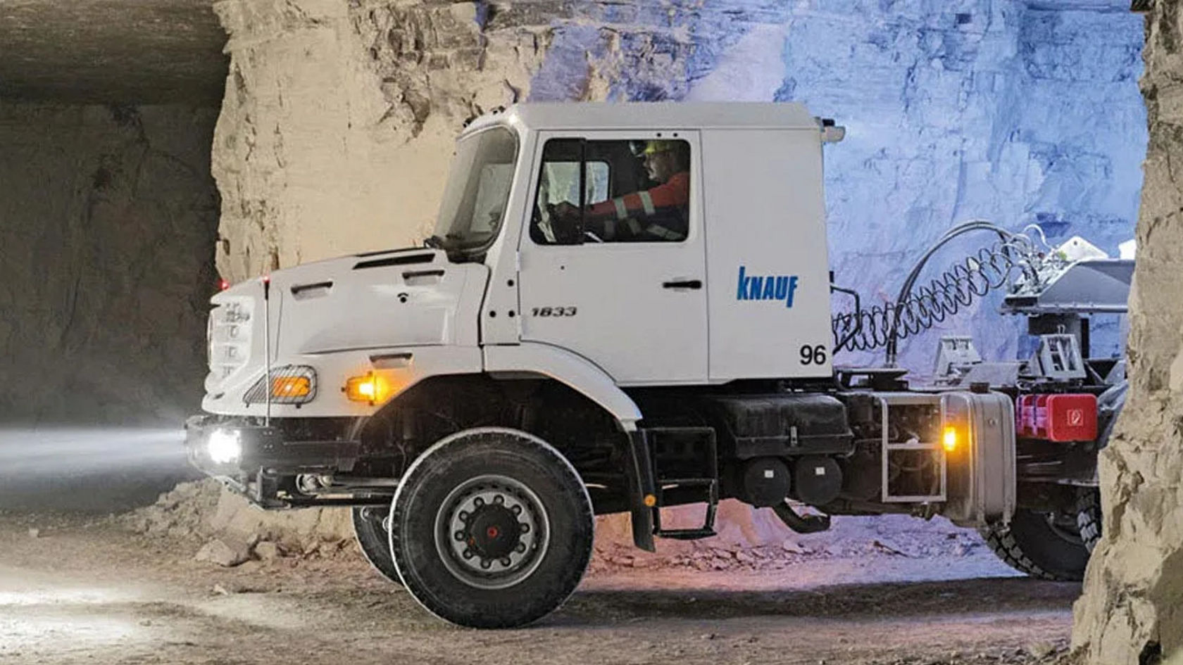 Un camion Mercedes‑Benz circule dans une grande carrière éclairée. Le camion a une cabine claire portant l'inscription « Knauf ». Il fait nuit et les phares sont allumés.