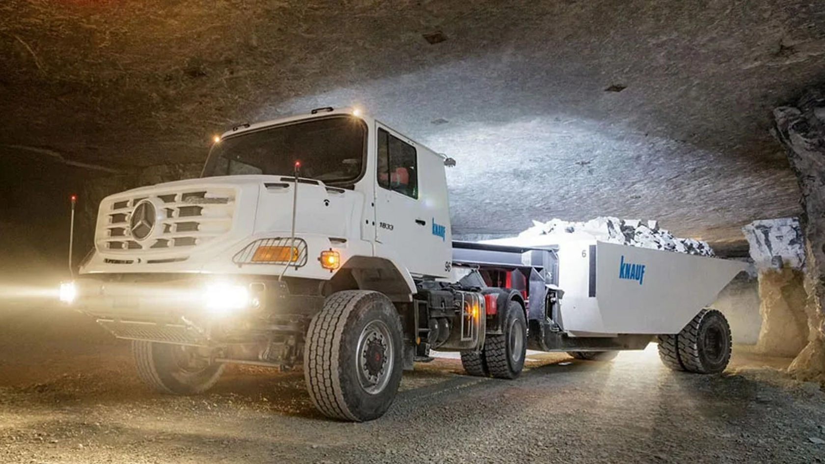 Un camion Mercedes-Benz traverse une galerie sombre et tire une grande remorque chargée de roches. Les phares avant éclairent la voie devant lui.