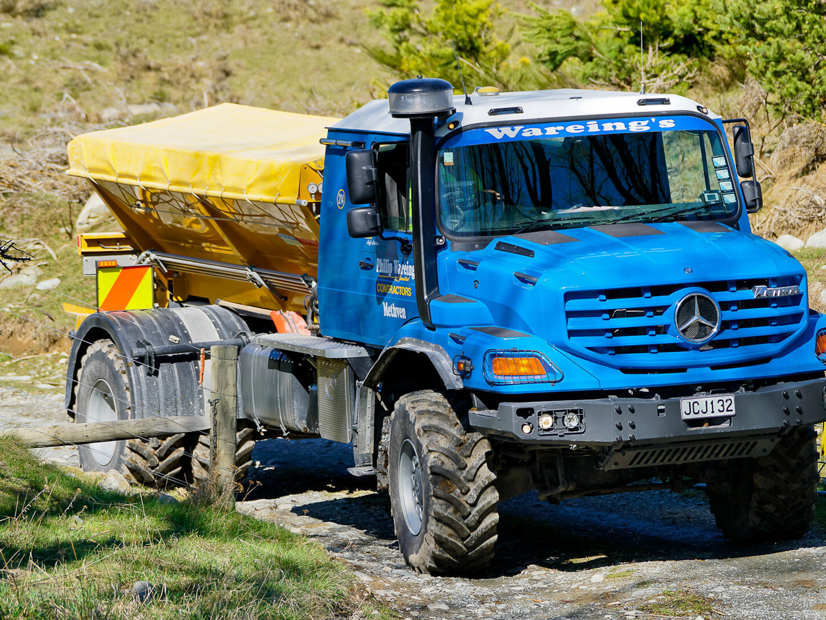 Un camion Mercedes‑Benz bleu tractant une semi‑remorque jaune circule sur une route étroite. Les pneus sont adaptés aux terrains accidentés, ce qui souligne son aptitude tout‑terrain.