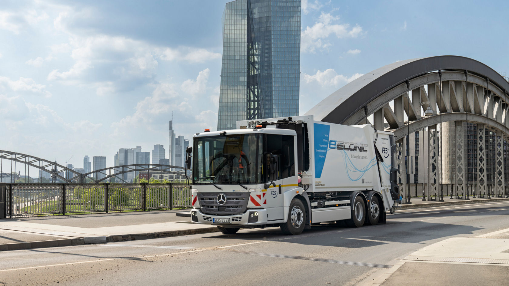Der Mercedes-Benz Econic Müllwagen steht auf einer Brücke mit einer modernen Stadtlandschaft im Hintergrund, während ein Arbeiter mit der Entleerung von Mülltonnen beschäftigt ist.
