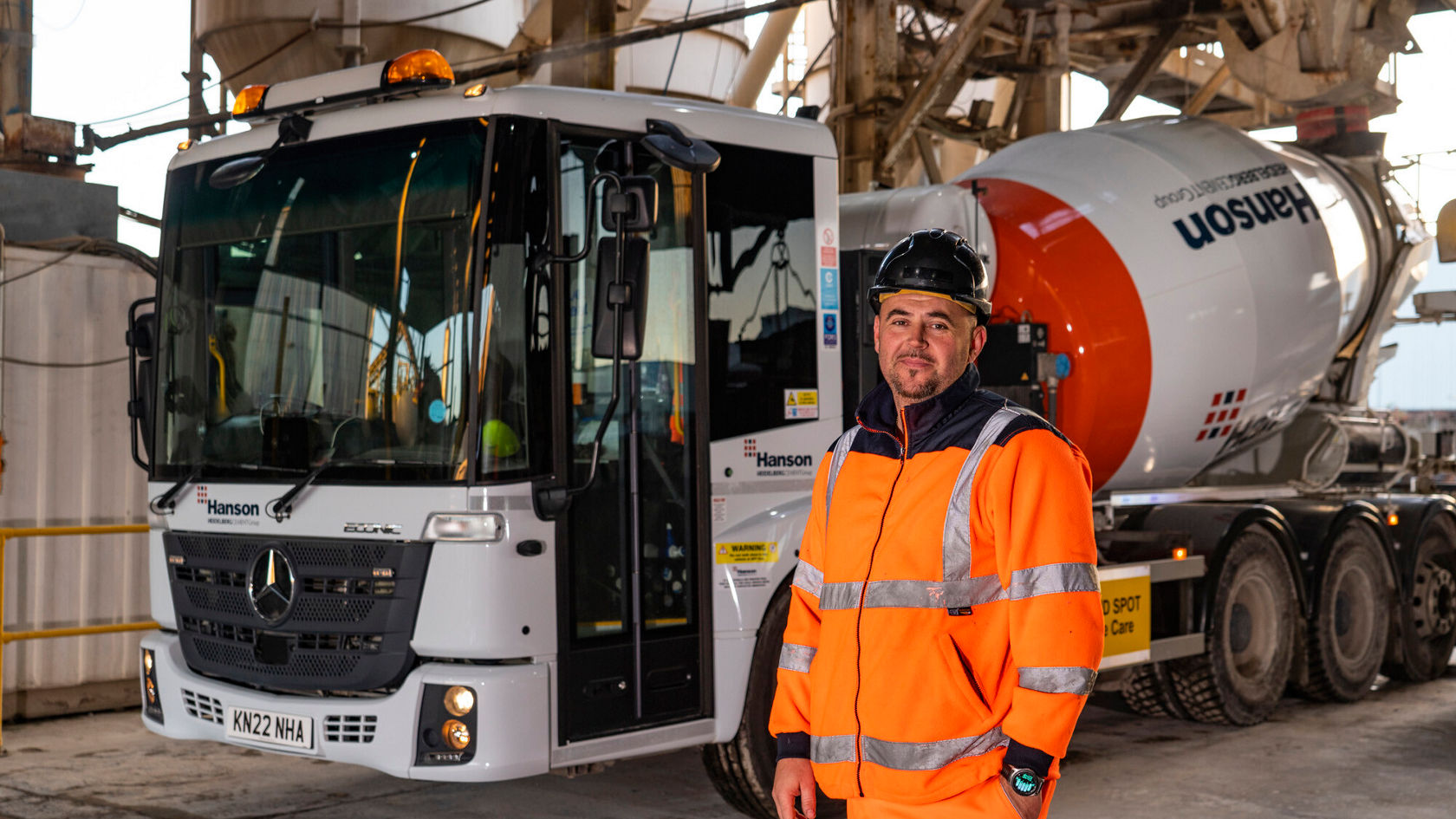 Ein Arbeiter in orangefarbener Sicherheitskleidung steht neben einem Mercedes-Benz Econic Betonmischer von Hanson in einer industriellen Umgebung, bereit für den Einsatz.