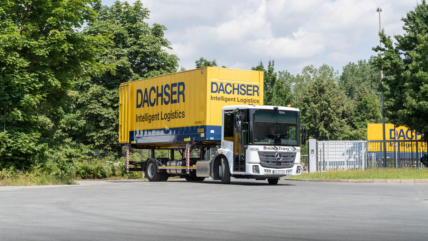 Ein Mercedes-Benz Econic-Lkw fährt durch das Tor eines Logistikzentrums, beladen mit einem gelben Dachser-Container, umgeben von Bäumen und einer Lagerhalle.