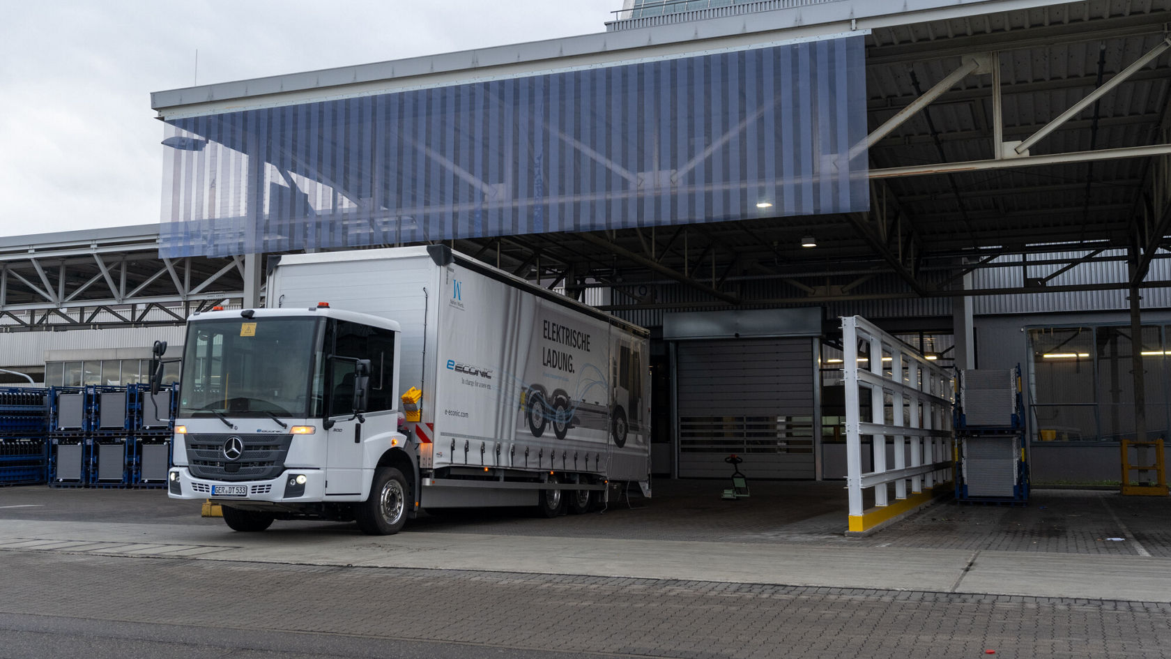 A Mercedes-Benz eEconic truck stands in front of a warehouse, equipped with a trailer. The truck is white with a large company logo and information about electric vehicles.