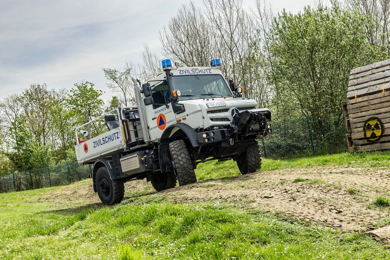 A Mercedes-Benz Unimog marked "CIVIL PROTECTION" stands on an unpaved track surrounded by green areas, equipped with a blue light and off-road tires.