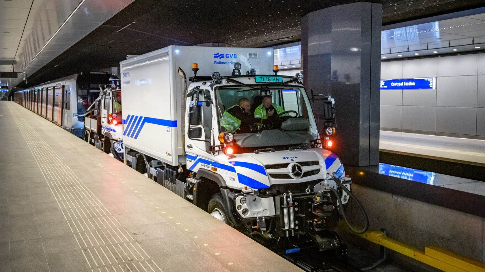 Mercedes-Benz Unimog steht an einem U-Bahnhof, während zwei Mitarbeiter im Fahrerhaus arbeiten. Der Unimog hat eine spezielle Ausstattung für den Einsatz im öffentlichen Verkehr.