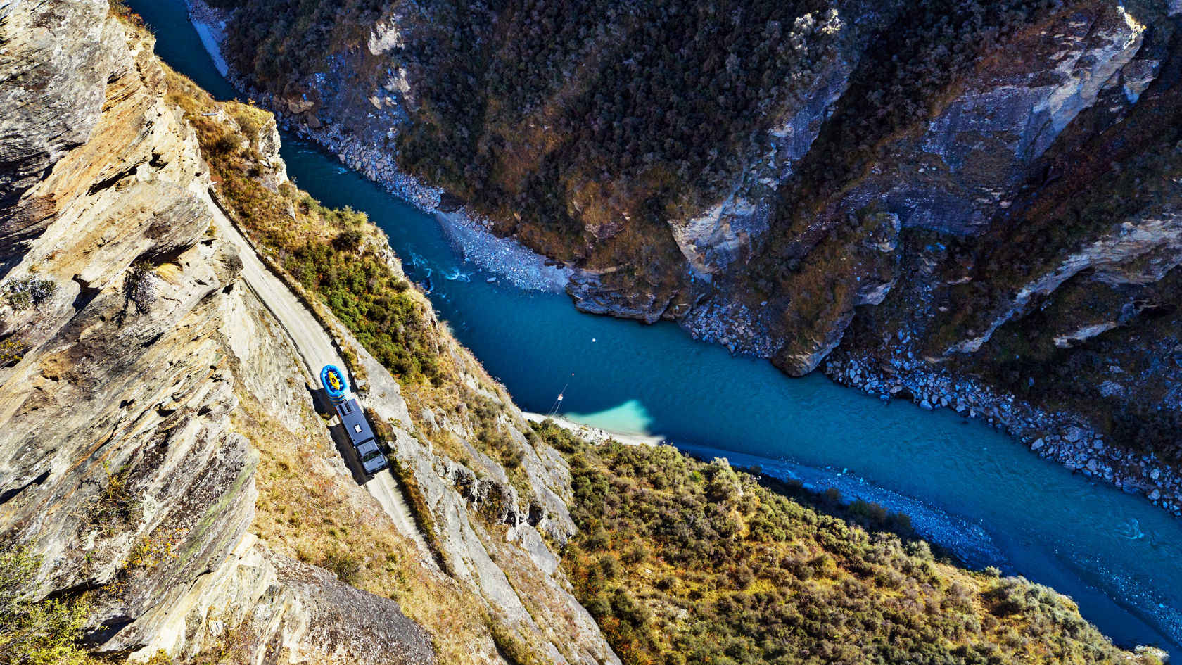 Ein Mercedes-Benz Unimog fährt aus der Vogelperspektive zu sehen auf einer Gebirgsstraße mit einem Fluss und felsigen Klippen im Hintergrund.
