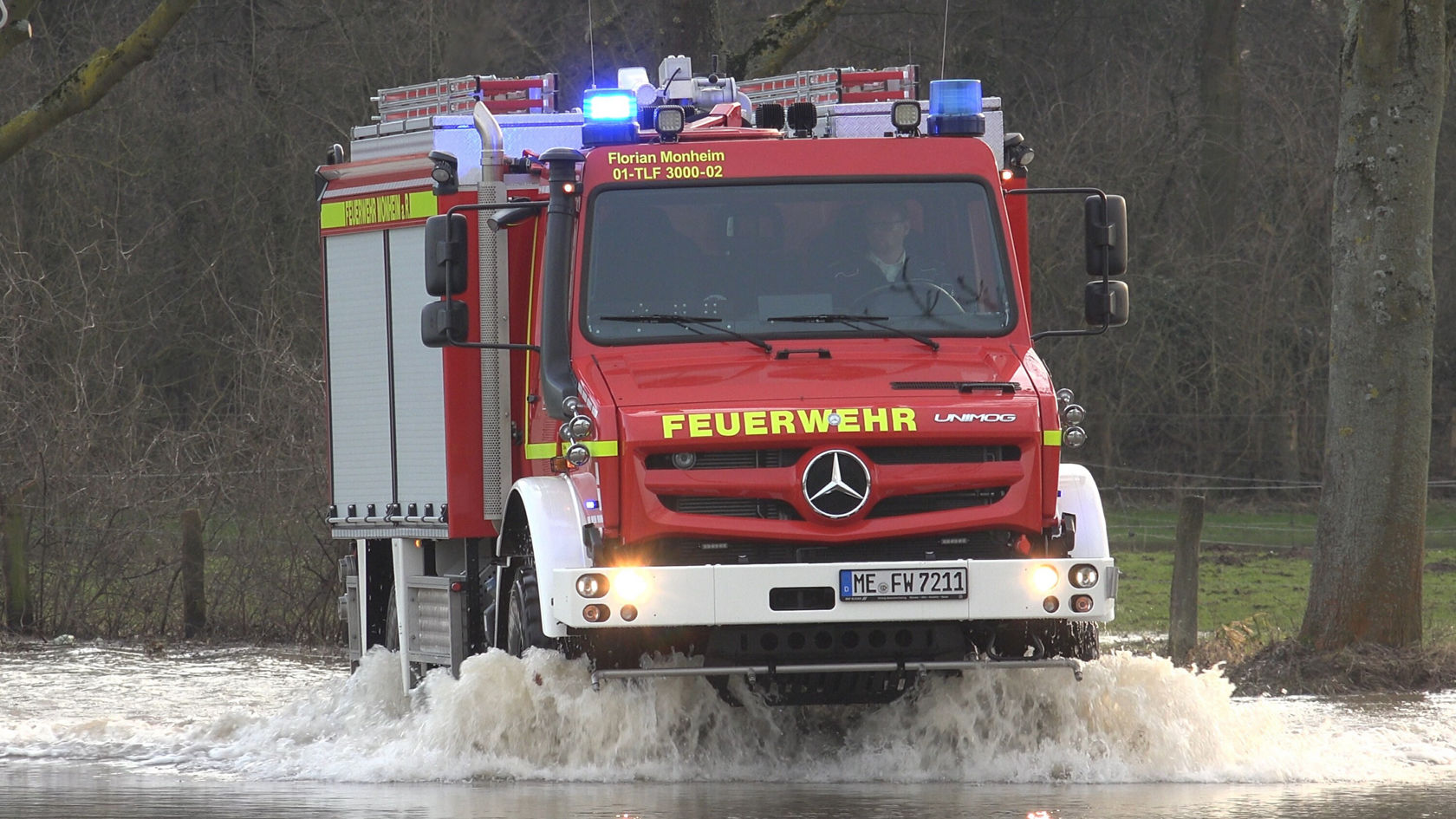 Un Unimog Mercedes‑Benz rouge, véhicule de pompiers, traverse une zone inondée.