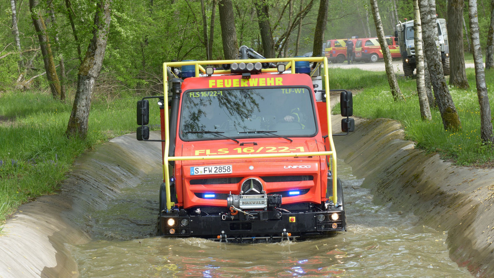 Un Unimog rouge des pompiers traverse un fossé rempli d'eau, le gyrophare allumé.