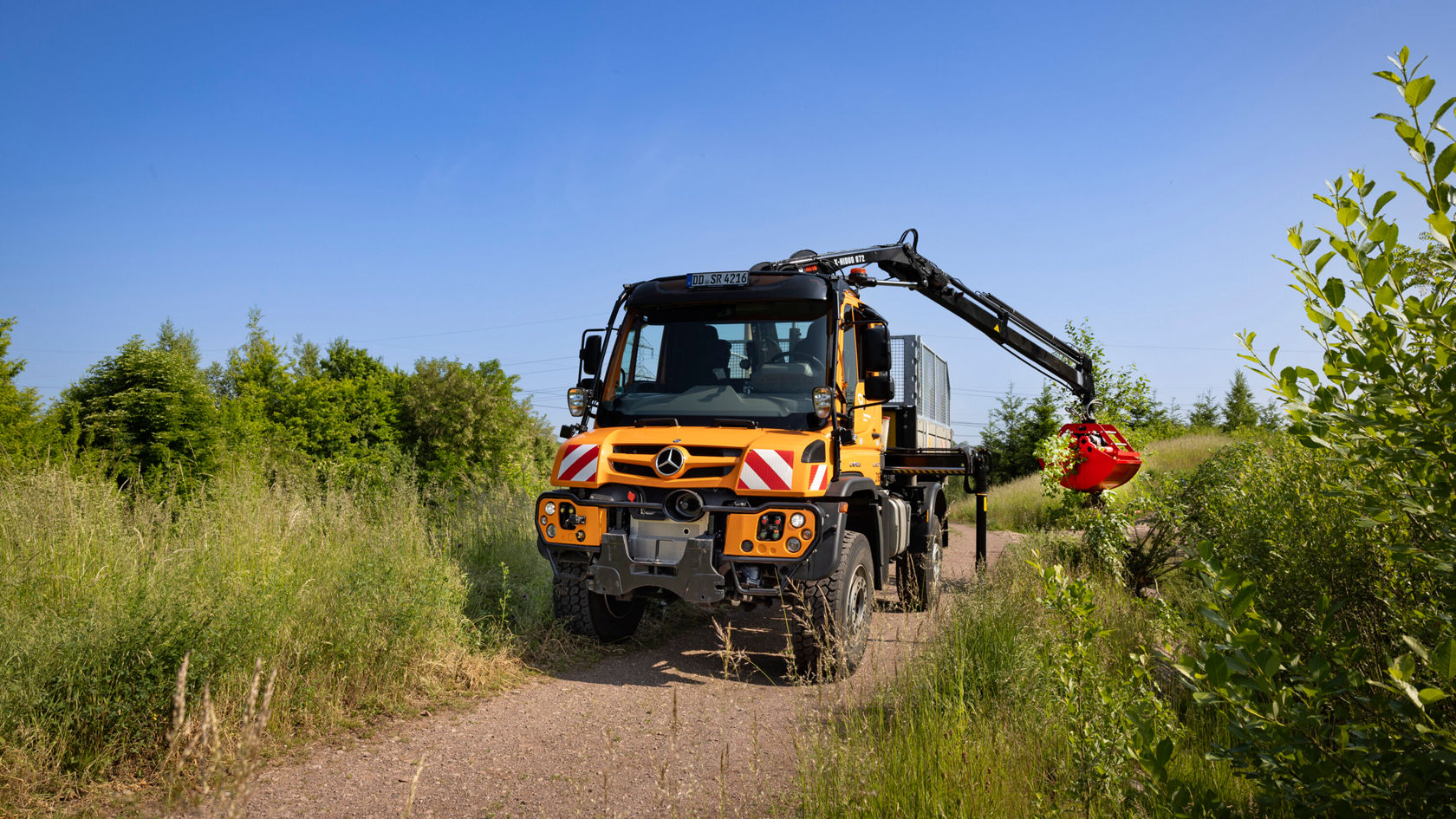 Ein Mercedes-Benz Unimog mit Kran hebt einen Baumstamm an, umgeben von Grünpflanzen und Bäumen auf einem unbefestigten Weg.