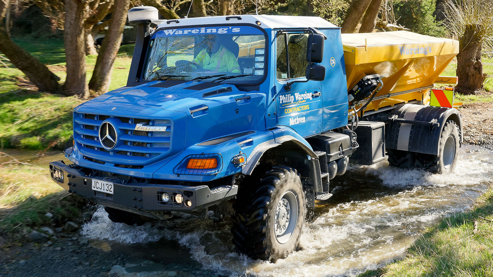 Un camion Mercedes‑Benz bleu traverse un petit ruisseau en transportant du matériel. Des gouttes d'eau jaillissent et deux personnes sont visibles à l'intérieur du véhicule.