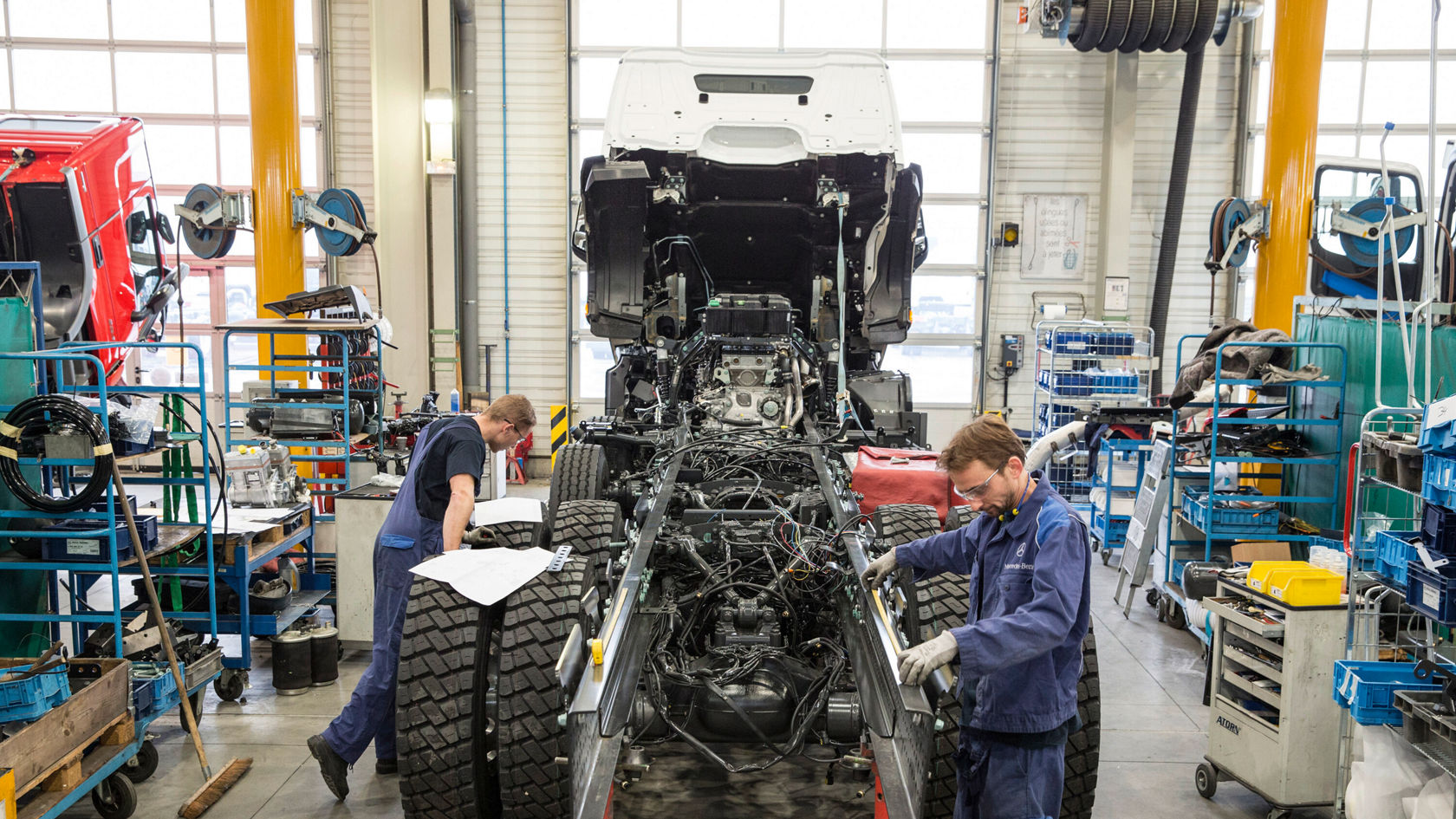 Zwei Mechaniker arbeiten in einer Werkstatt an einem Mercedes-Benz Lkw, während sie an der Technik und den Reifen des Fahrzeugs Hand anlegen.