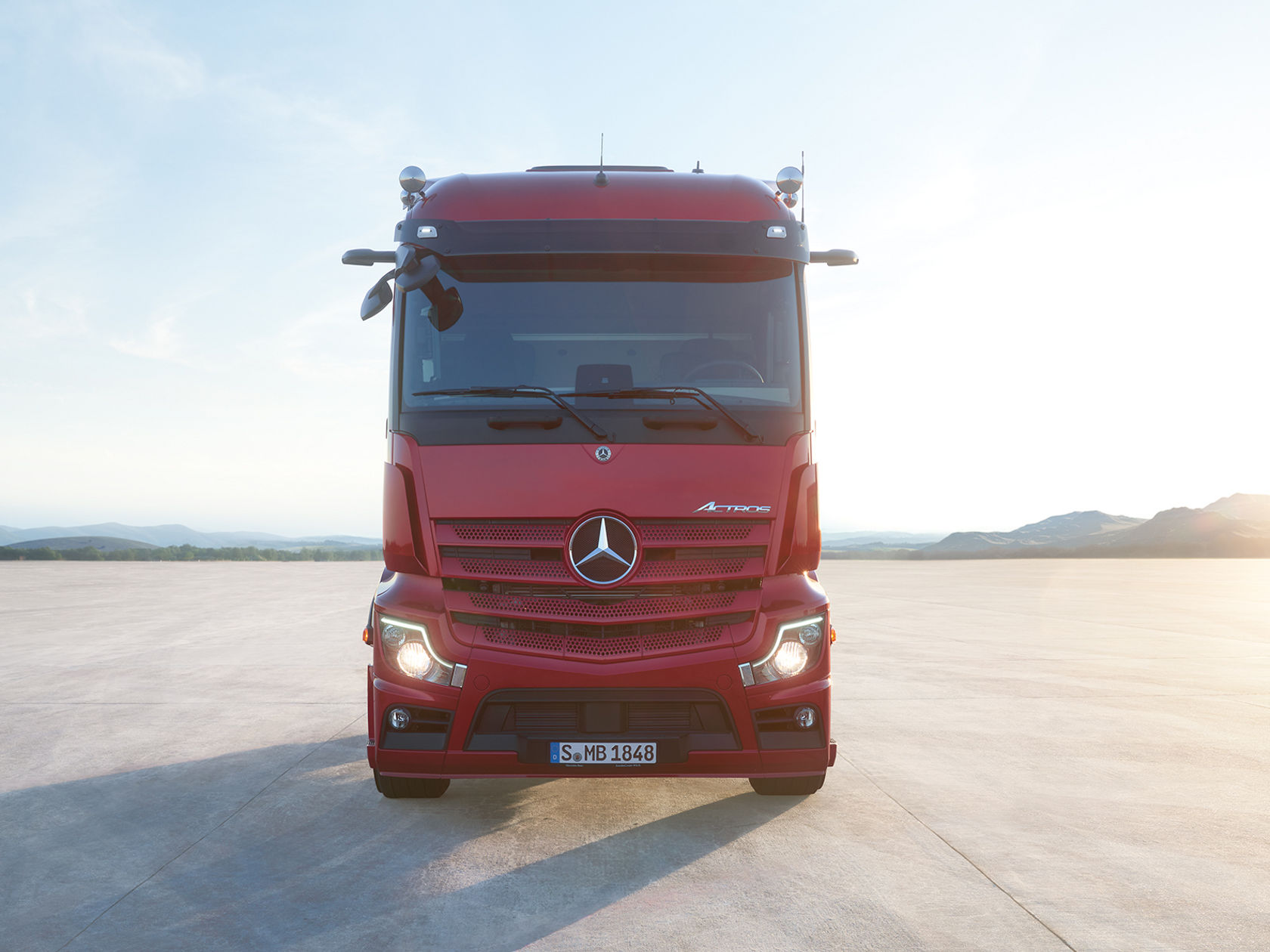 Ein roter Mercedes-Benz Actros LKW steht frontal auf einer weiten, hellen Fläche mit einem langen Schatten. Im Hintergrund sind Berge unter einem klaren Himmel zu sehen.
