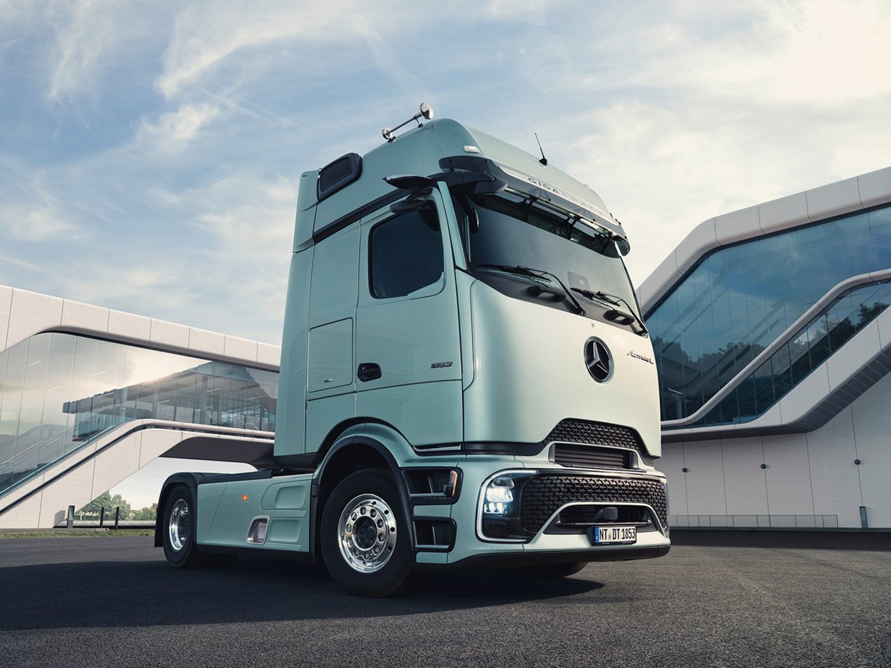 Ein mintgrüner Mercedes-Benz Actros L Lkw steht draußen vor einem modernen Gebäude mit Glasfassade und geschwungenen weißen Elementen unter einem hellblauen Himmel.