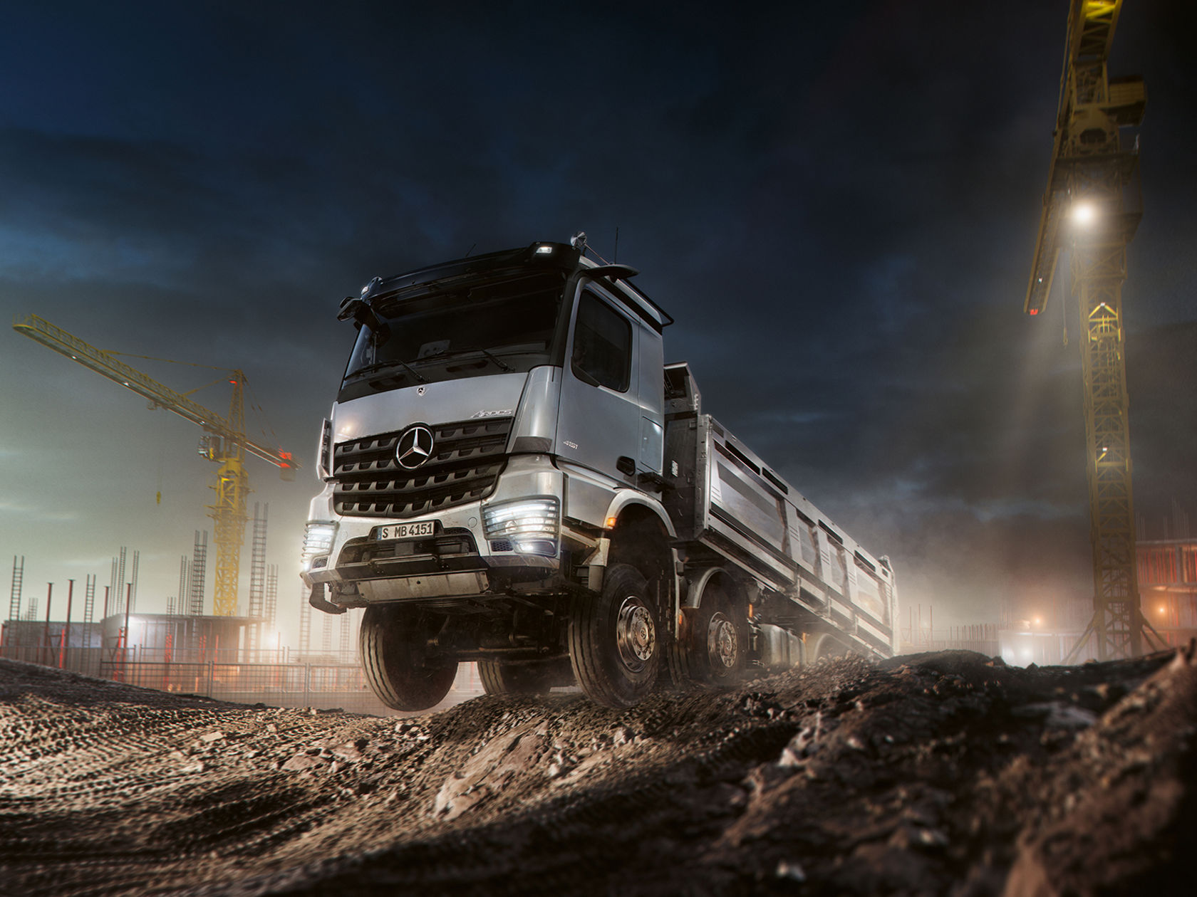 Ein silbergrauer Mercedes-Benz Arocs LKW fährt in der Dämmerung oder Nacht über eine unebene Baustelle. Im Hintergrund sind Kräne und Baustrukturen zu sehen, beleuchtet von Scheinwerfern und dem Licht des LKWs.