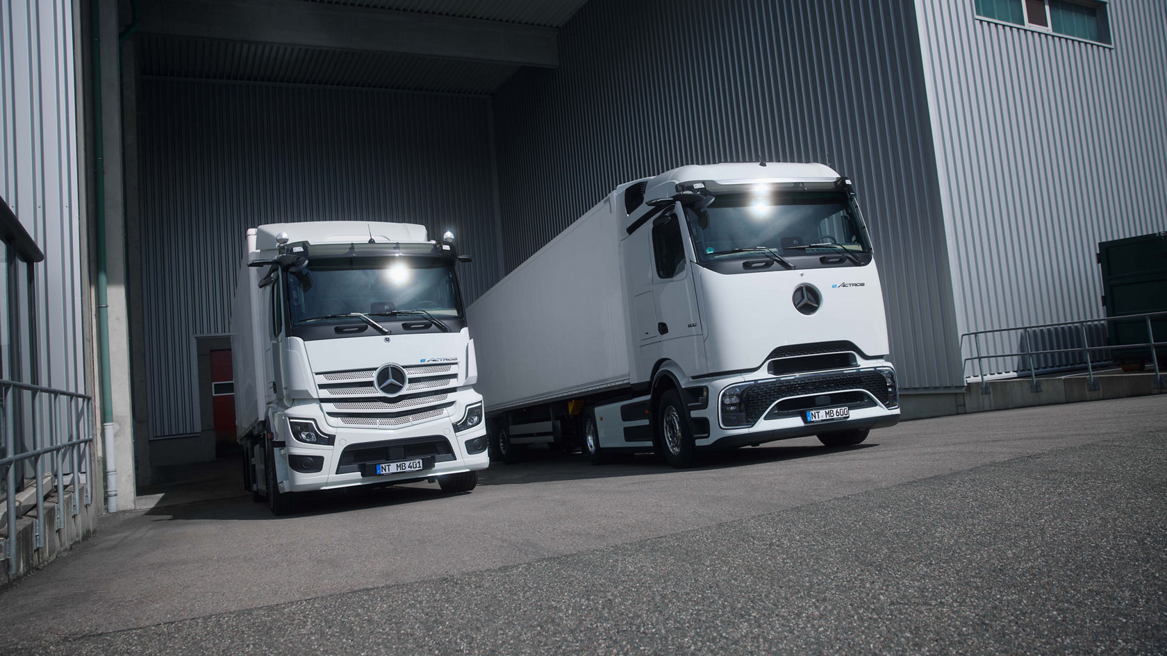 Zwei weiße eActros von Mercedes-Benz Trucks, einer mit ProCabin und einer mit bewährter Kabine, stehen frontal nebeneinander auf einer asphaltierten Fläche vor einer Betonwand. Beide Lkw haben ihre Scheinwerfer an und im Hintergrund sind Bäume und ein modernes Gebäude zu sehen.