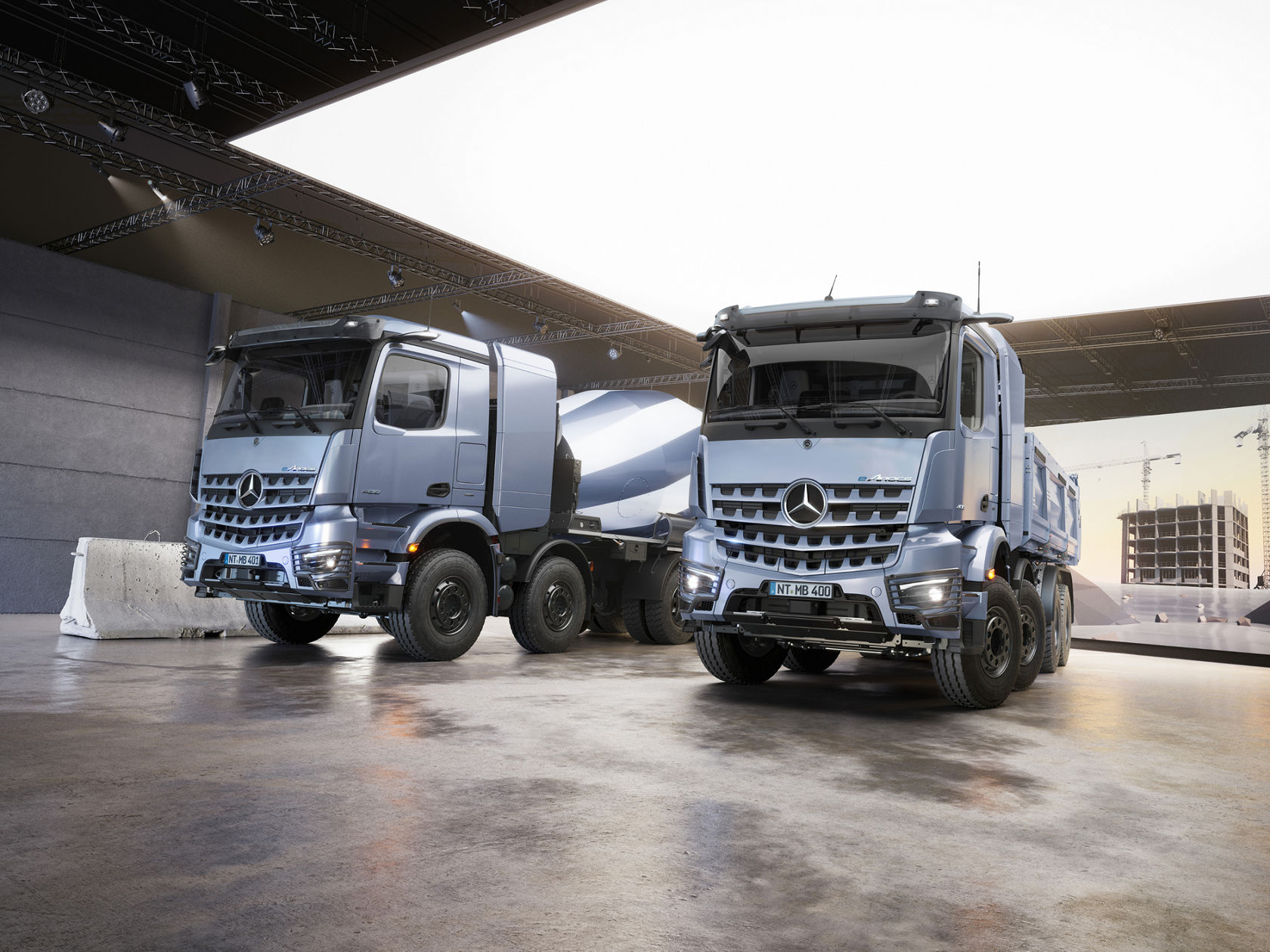 De afbeelding toont twee eArocs 400 in een grote hal met betonnen vloer en plafondverlichting. De eArocs 400 aan de rechterkant is een betonmixer, terwijl de eArocs 400 aan de linkerkant een kipper is. Op de achtergrond is een groot led-scherm te zien met een afbeelding van een bouwplaats.