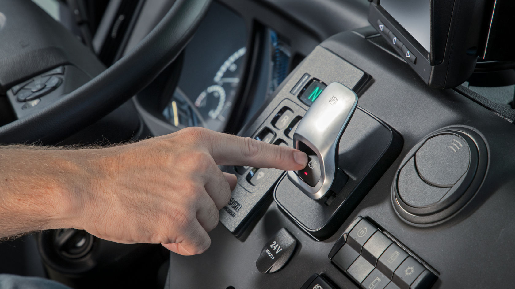 Een hand drukt op een knop op een elektronische versnellingshendel. De versnellingshendel bevindt zich op de middenconsole en is omgeven door andere bedieningselementen en displays.