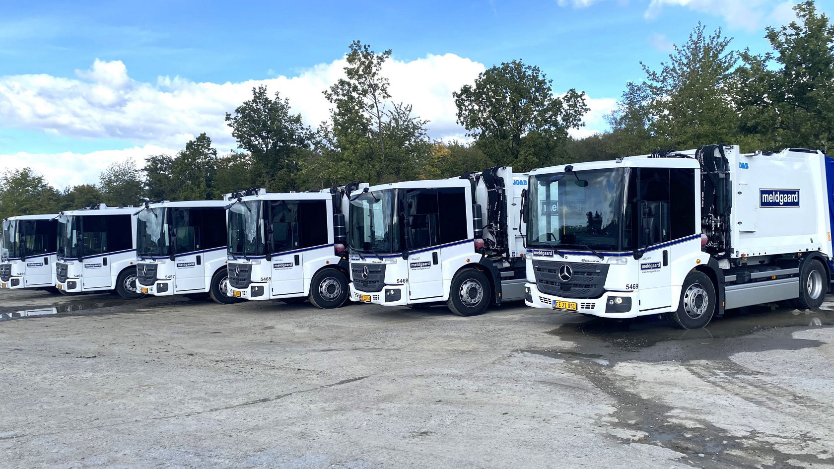 Fünf Mercedes-Benz Econic Müllfahrzeuge stehen nebeneinander auf einem Gelände, umgeben von Bäumen und unter einem bewölkten Himmel.