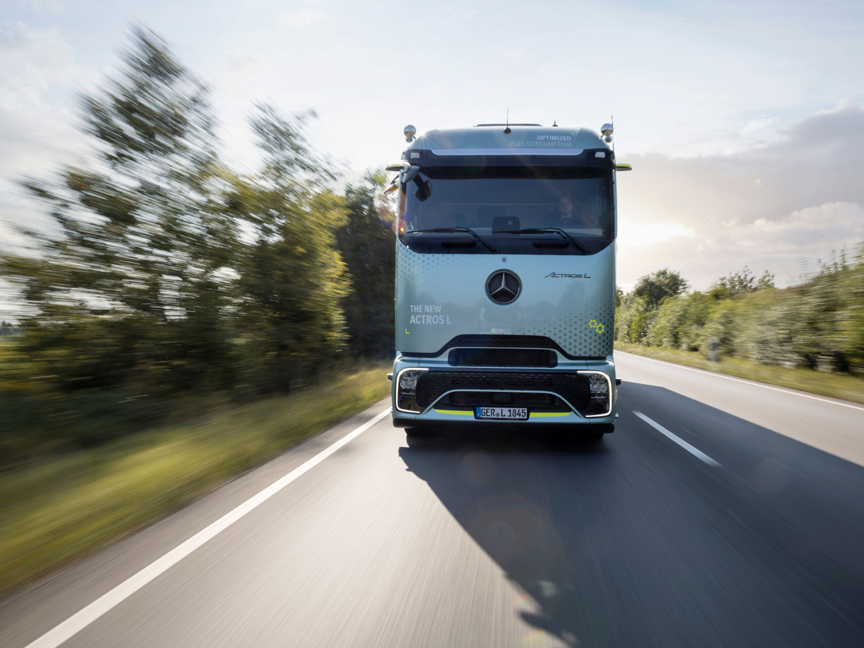 Een mintgroene Mercedes-Benz Actros L met ProCabin rijdt op een straat met bomen en wordt van voren getoond. De zon schijnt fel en werpt een lange schaduw van de vrachtwagen op het asfalt.