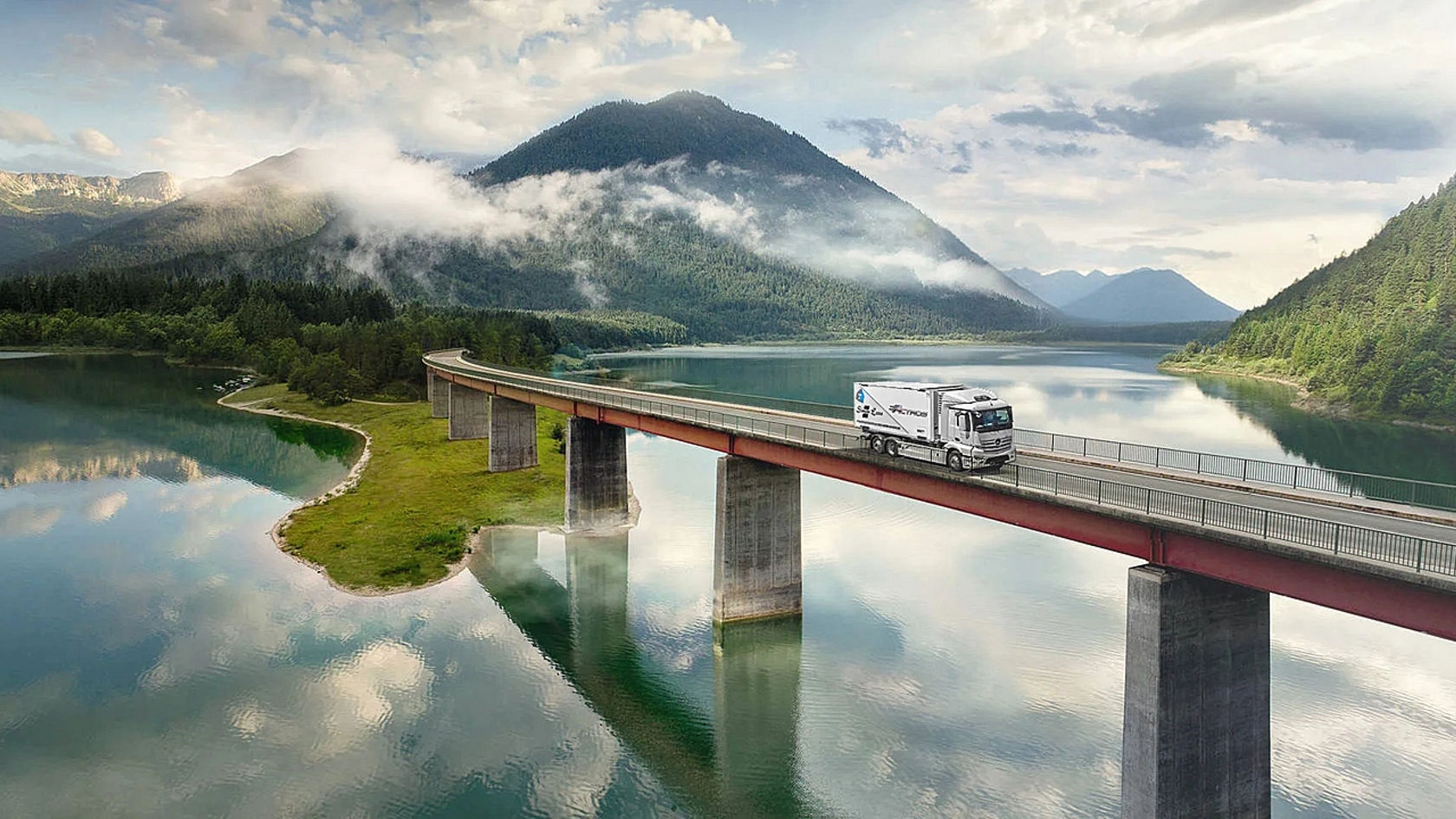 Der eActros 300/400 fährt über eine Brücke, die einen See in einer Berglandschaft überspannt.