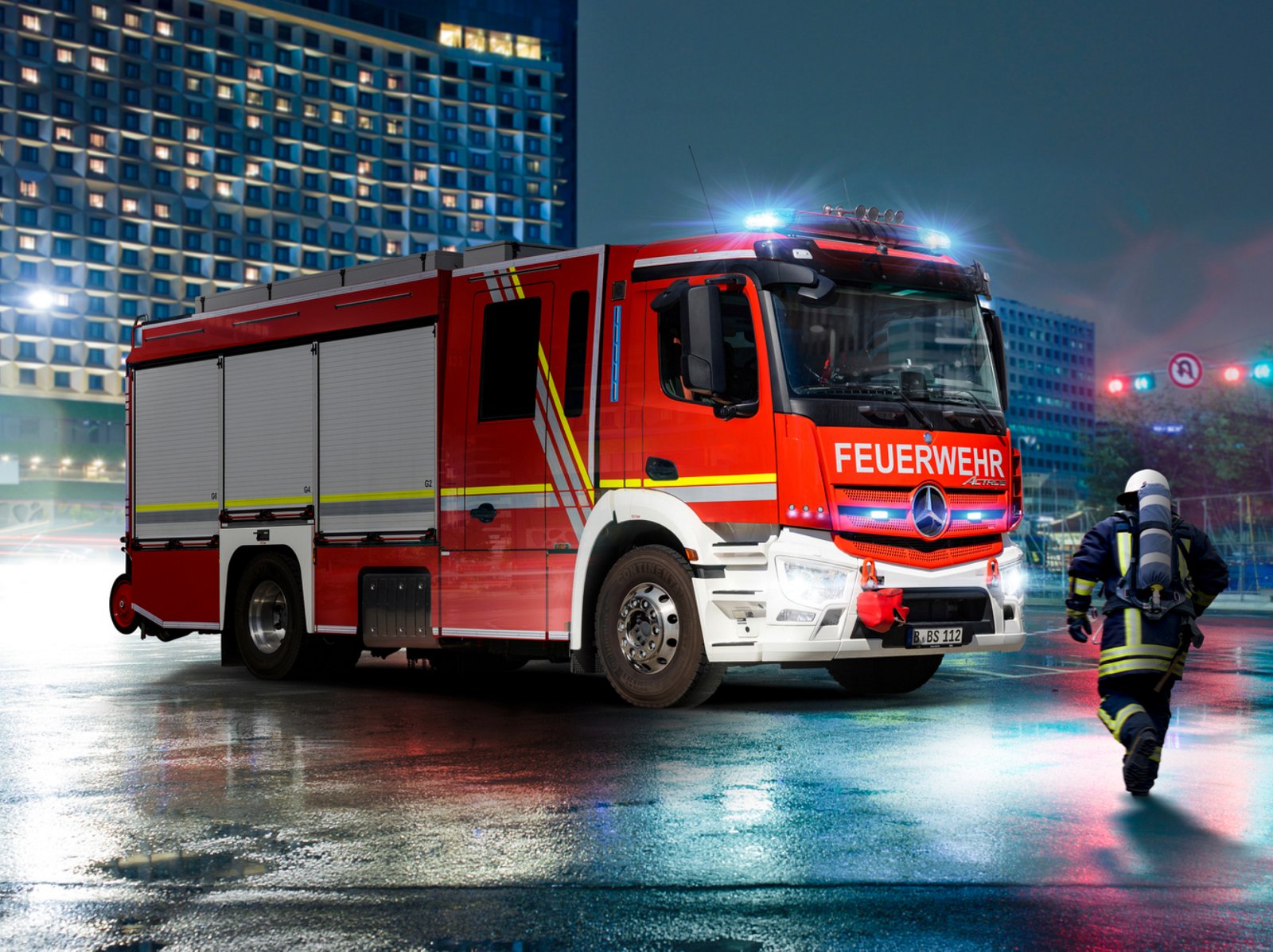 An Actros fire-fighting vehicle with its blue lights on stands on a wet road in the city. A firefighter in full gear is running towards the vehicle. In the background is a large building with illuminated windows. The scene seems dark and urgent.