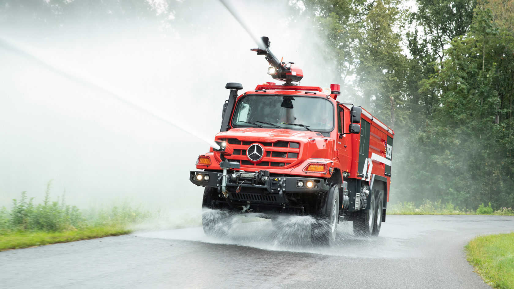  A Unimog airport fire-fighting vehicle driving on a road and using two water cannons to dispense large quantities of water or extinguishing foam. The first cannon is located on the vehicle roof, the second at the height of the headlights. The spray indicates a high speed.