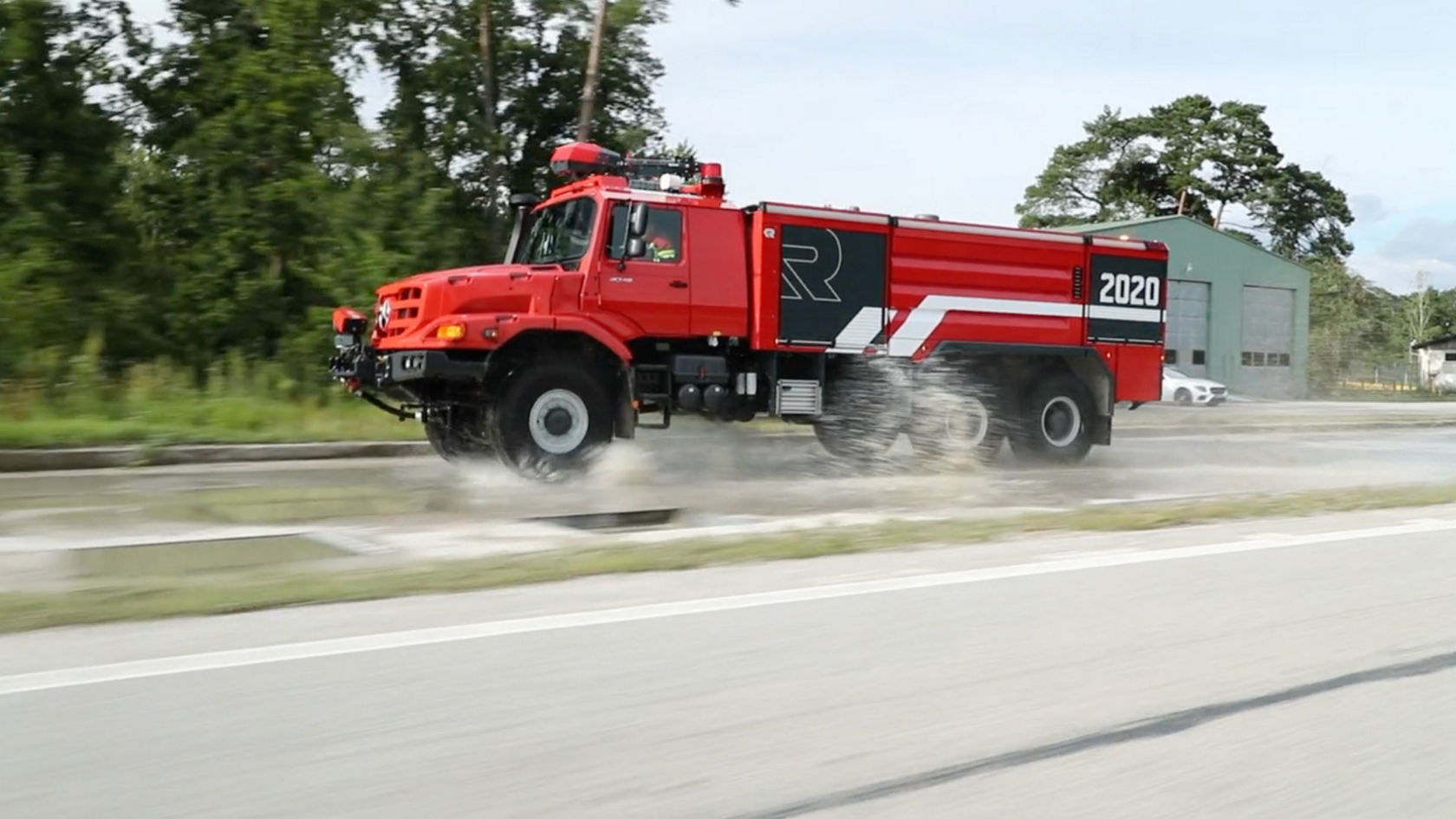 The image shows a red Unimog as an airport fire-fighting vehicle. The vehicle is rolling over a wet road. The body bears a large “R” and the year “2020”. A warehouse and trees can be seen in the background. 