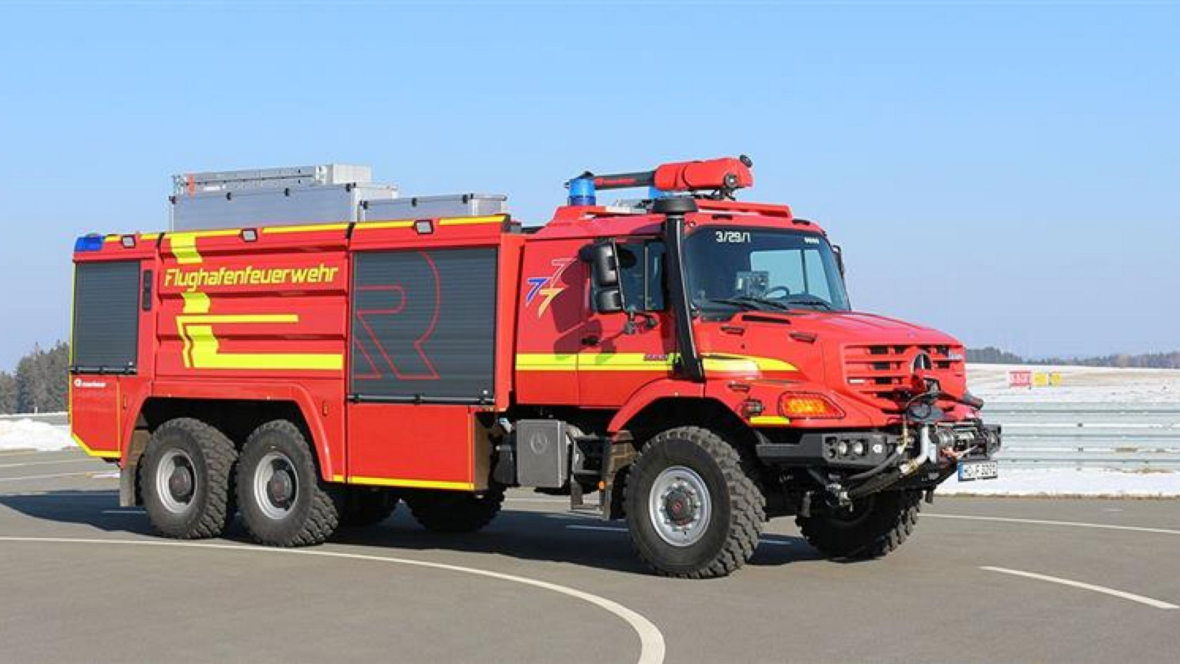 A large airport fire-fighting vehicle with the inscription “Flughafenfeuerwehr” standing in front of an airport. The Zetros features several axles and large tyres, which suggests high off-road capability and a large extinguishing agent capacity. An asphalt road and snow is visible in the background.