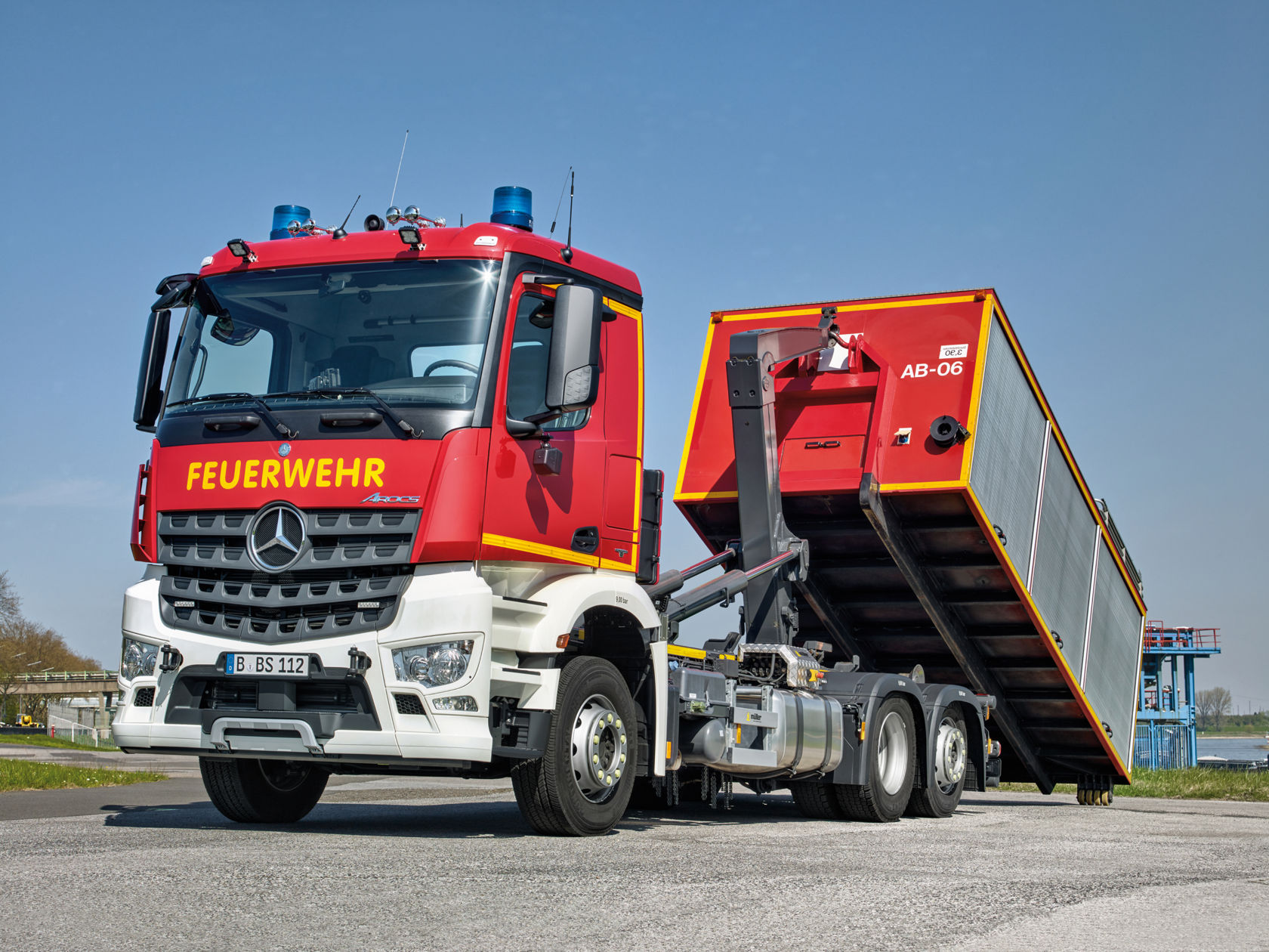 An Arocs fire-fighting vehicle is parked on an asphalt surface. The vehicle has a red and grey container body and says “AB-06”. Blue lights are visible on the roof of the vehicle. The sky is clear and blue. Trees and a bridge are visible in the background.
