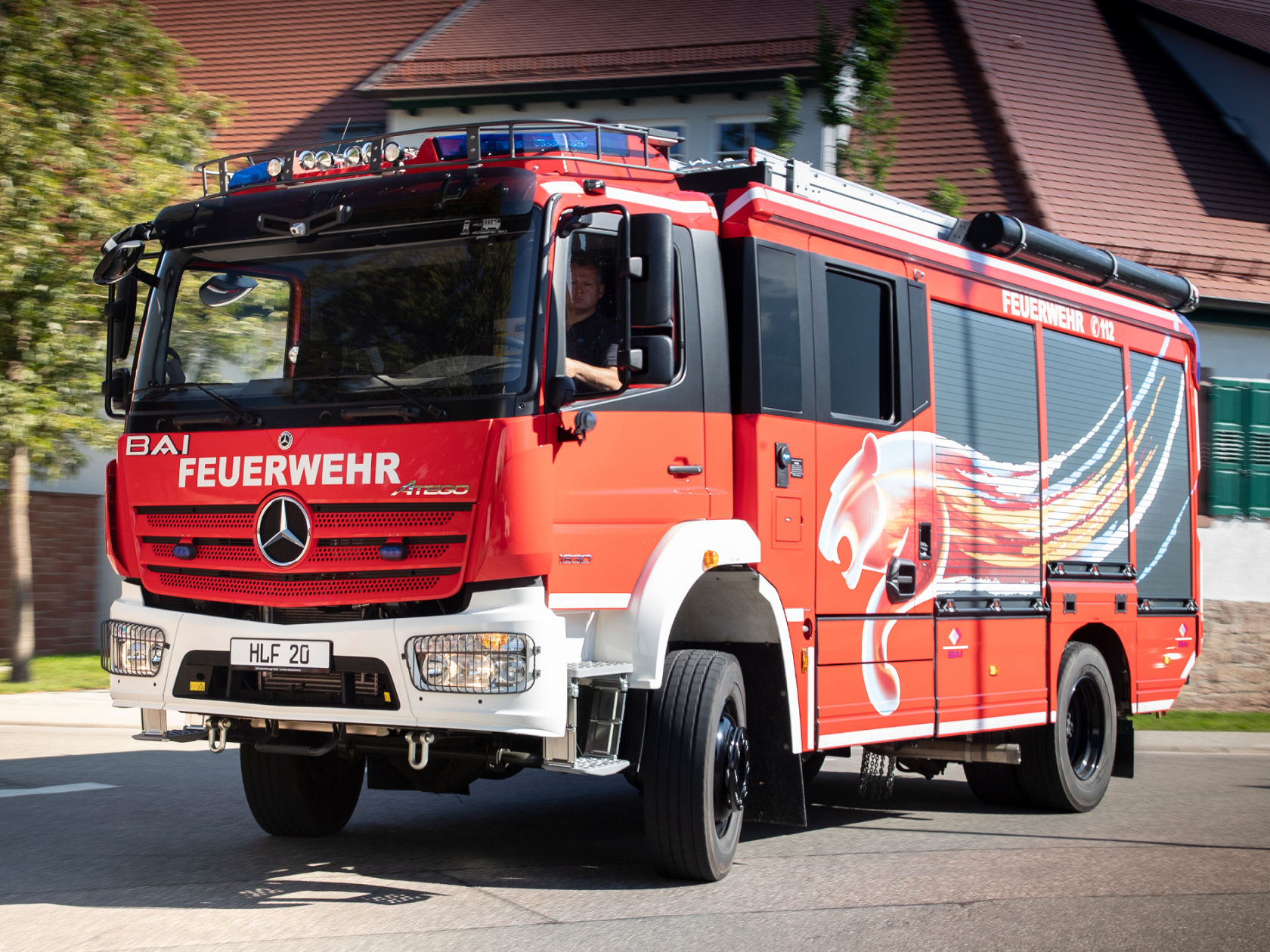 A red Atego fire-fighting vehicle with blue lights on drives on a road past a timbered house and trees. The front of the vehicle says “FEUERWEHR”.