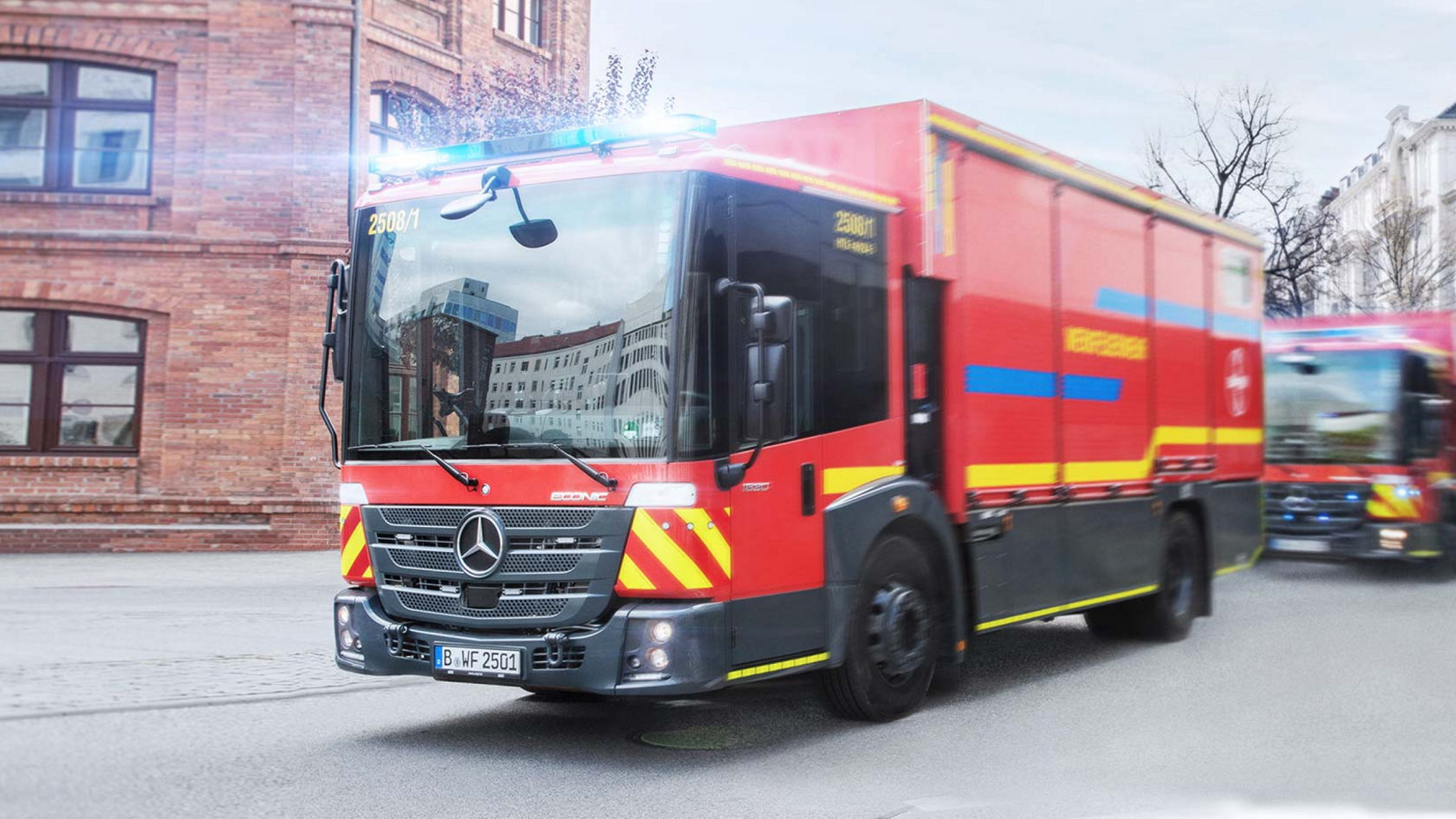 An Econic driving along a city road with blue lights on. The fire-fighting vehicle has a low cab and a large panoramic windscreen. Another vehicle is following behind. There is a large brick building in the background. 