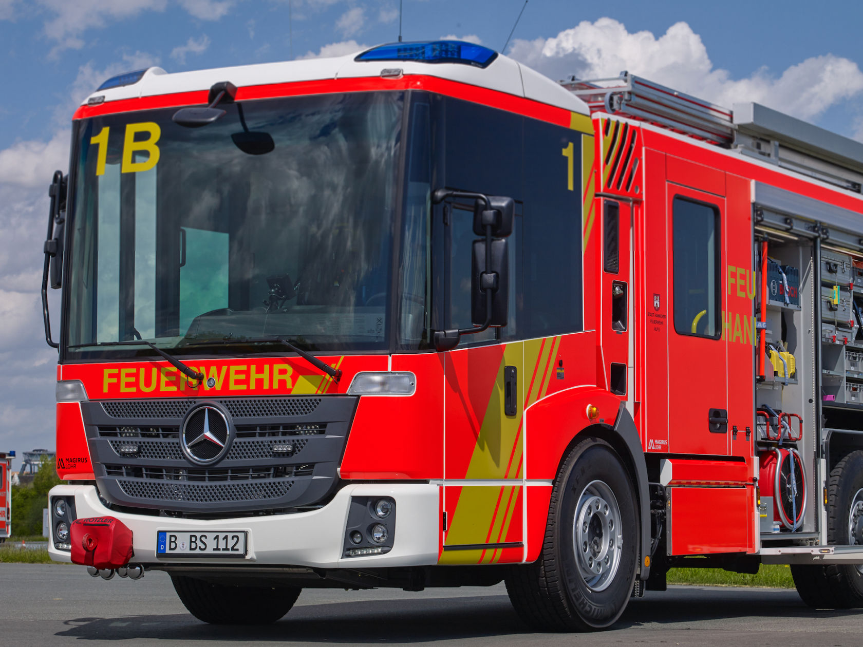 An Econic drives along a city road with blue lights on. The fire-fighting vehicle has a low cab and a large panoramic windscreen. Another vehicle is following behind. There is a large brick building in the background. 