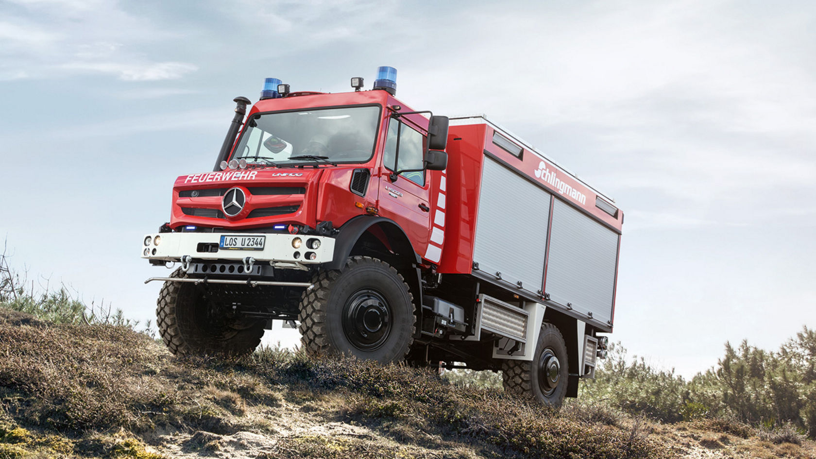 A red fire service Unimog driving up a steep, sandy slope with blue lights on. The off-road vehicle is equipped with heavy-duty tyres and a special body, presumably for fire-fighting operations. “Feuerwehr” and “Schlingmann” lettering indicate its intended use and the bodybuilder.