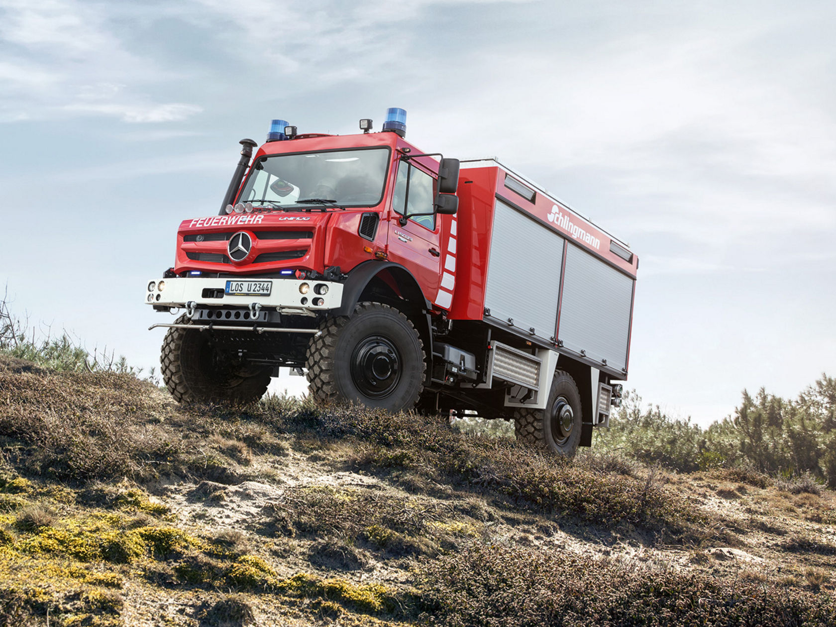 A red fire service Unimog driving up a steep, sandy slope with blue lights on. The off-road vehicle is equipped with heavy-duty tyres and a special body, presumably for fire-fighting operations. “Feuerwehr” (<i>Fire</i> <i>service</i>) and “Schlingmann” indicate its intended use and the bodybuilder, respectively.