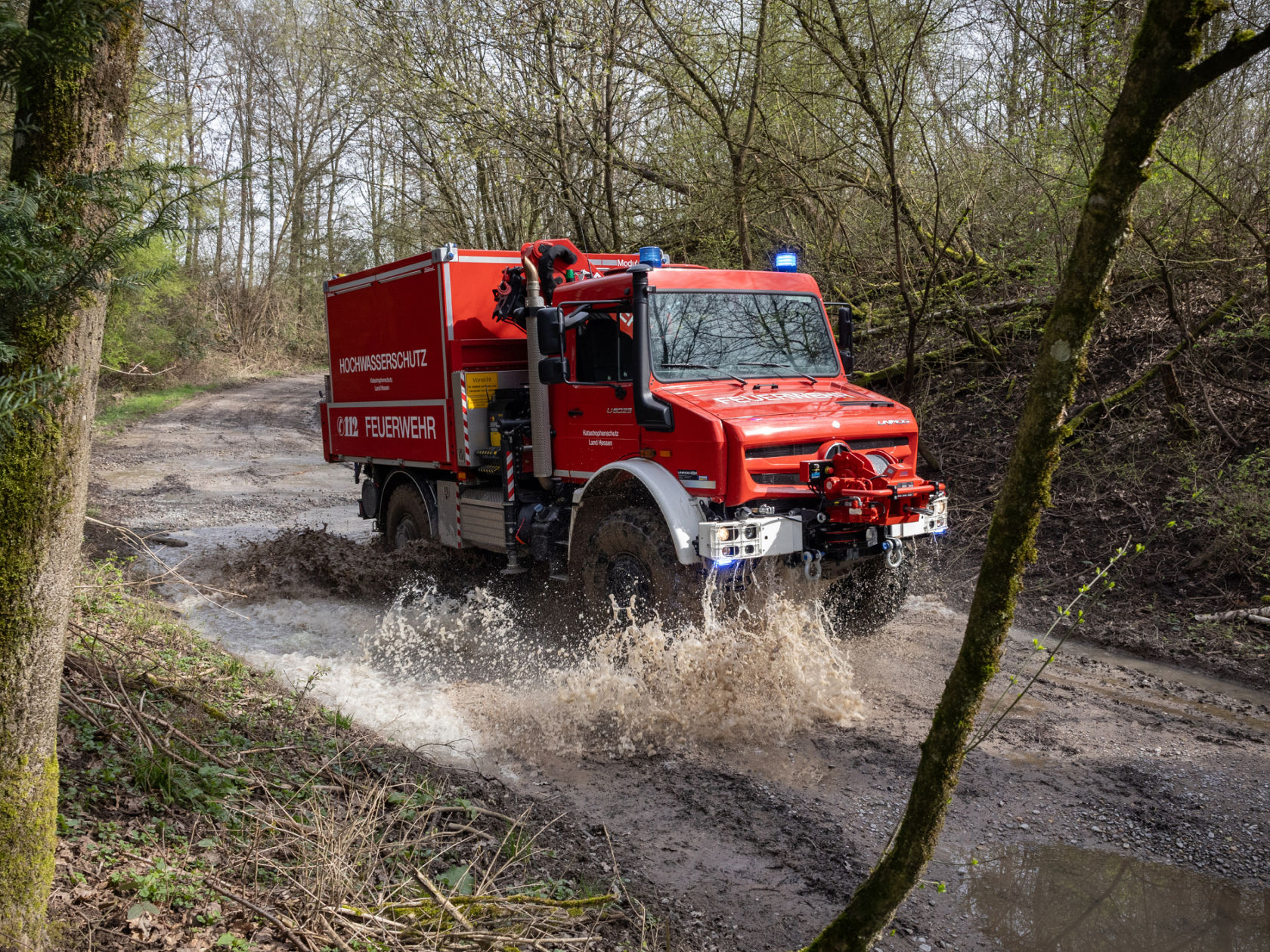 A fire service Unimog driving through a large puddle on a muddy forest road. The vehicle splashes up large amounts of mud and water. “Hochwasserschutz” lettering indicates its intended use.