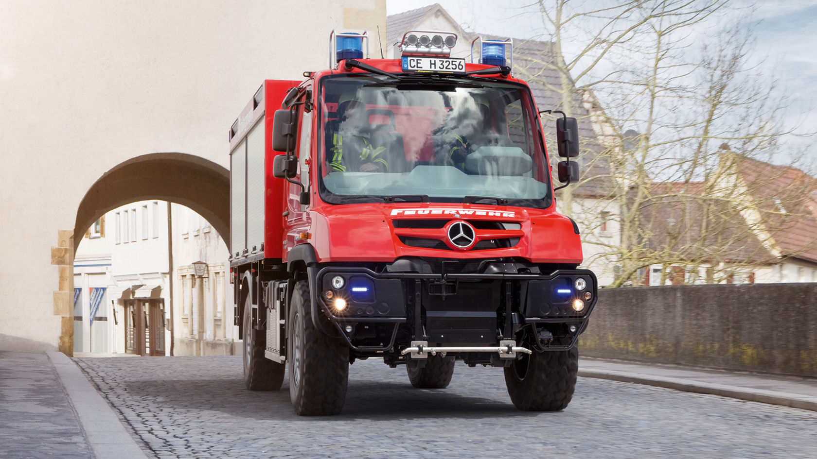 A red Unimog with blue lights on driving over a paved path through a city gate. The ground is cobbled. Two firefighters sitting in the vehicle