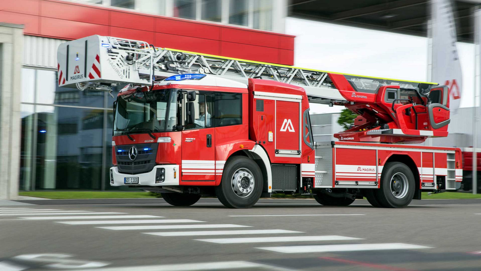 A red and white fire service Econic with a turntable ladder driving across a pedestrian crossing. The vehicle has a long extendable ladder arm. In the background, blurred industrial buildings indicate the speed of the Econic.