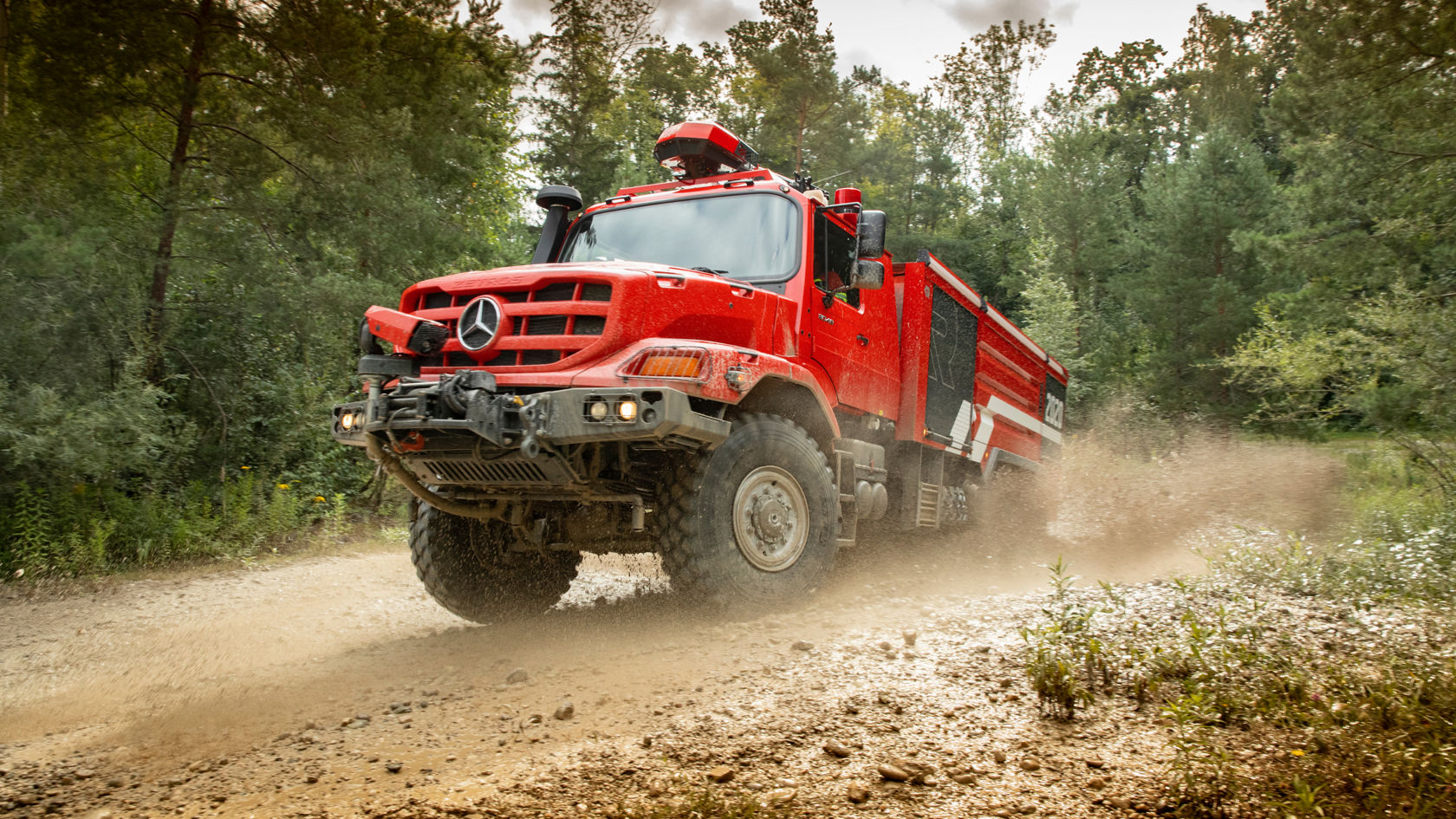 A red fire service Zetro fire-fighting tanker passing quickly over a wet field path, swirling up soil. The large tyres and high ground clearance illustrate the off-road capability of the vehicle. A firefighter at the wheel is visible through the windscreen. 