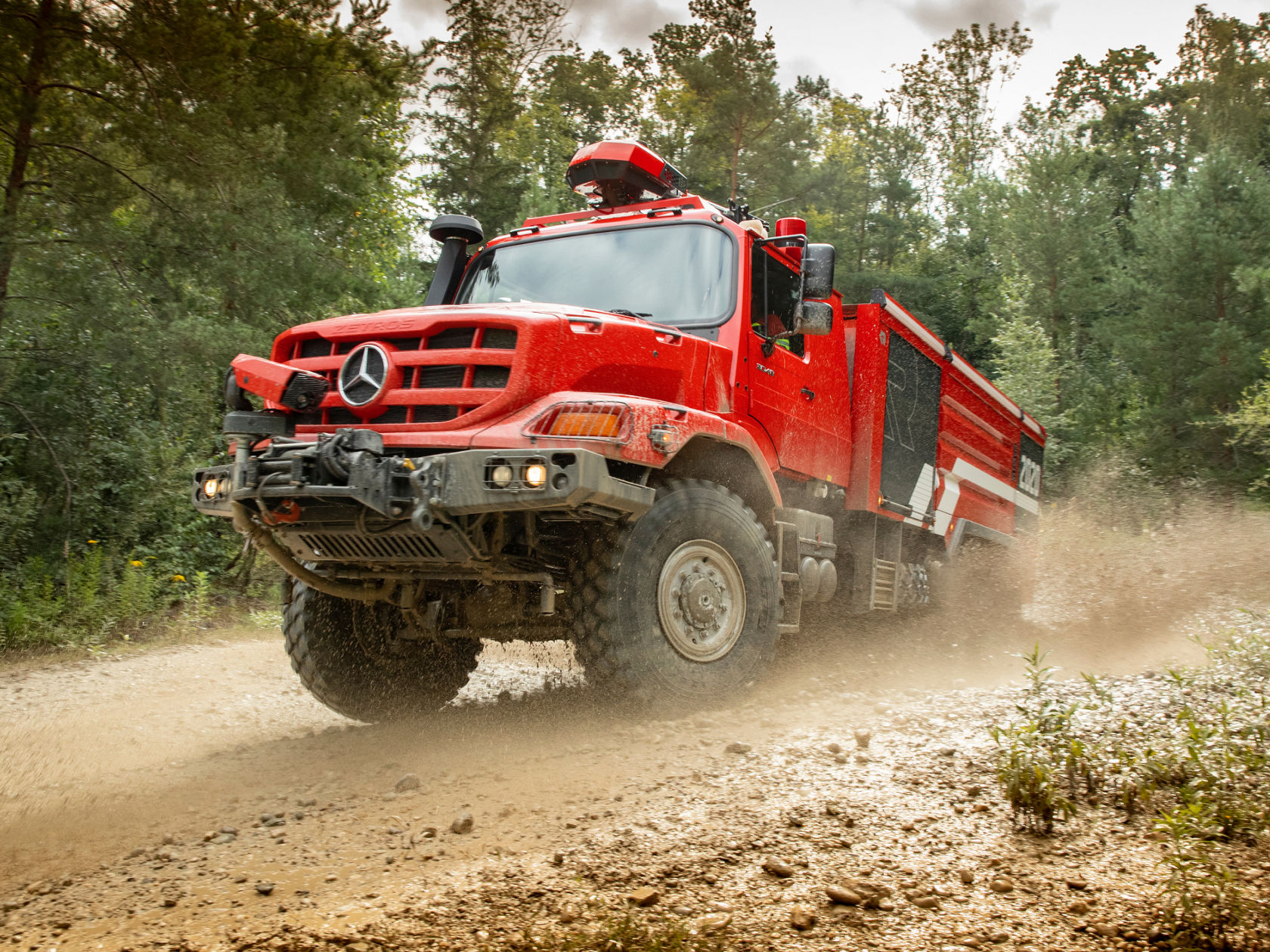 A red fire service Zetros fire-fighting tanker passing quickly over a wet field path, swirling up soil. The large tyres and high ground clearance illustrate the off-road capability of the vehicle. A firefighter at the wheel is visible through the windscreen. 