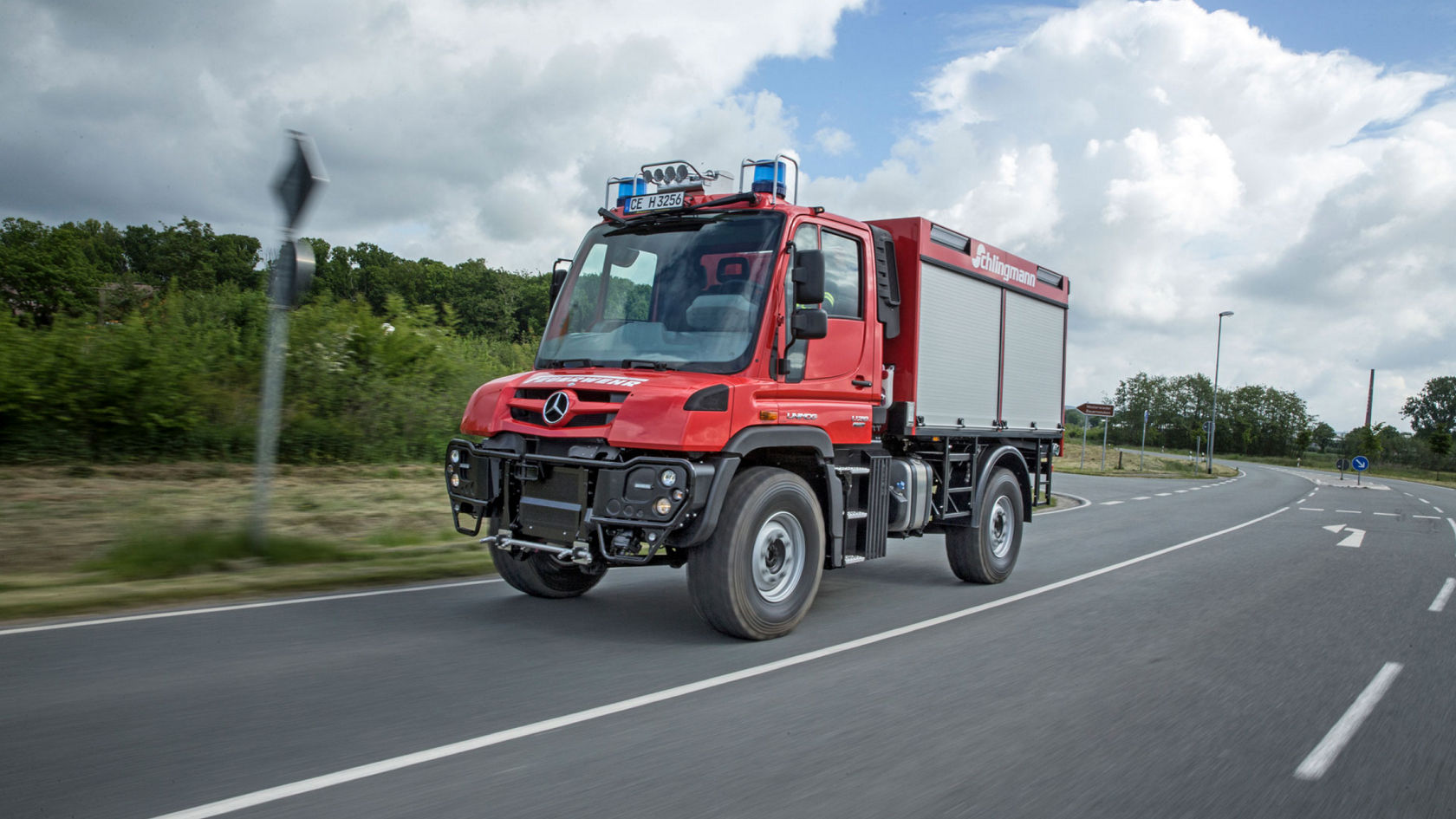 A Unimog driving along a road with blue lights on. The vehicle is painted red and has a fire service body. Trees and bushes can be seen in the background. 