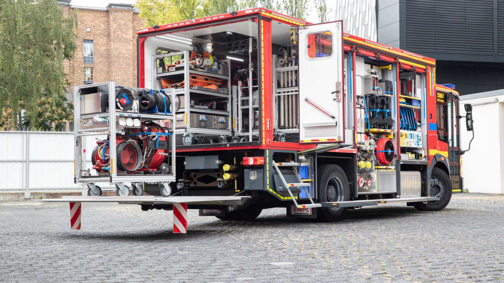 A red fire service Econic with open roller shutters standing on a cobbled square. The body of the equipment truck shows a variety of equipment items and devices that can be accessed via an extendable loading surface. 