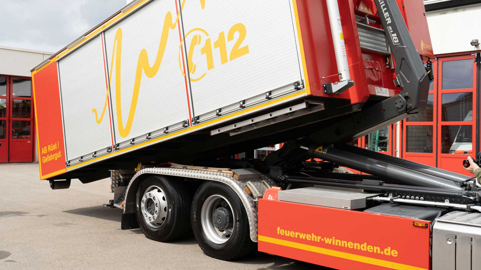 The image shows a red truck in front of the fire service garage. A tipper system vehicle with a fire-fighting container raises its loading surface. The yellow “feuerwehr-winnenenden.de” lettering is visible. 