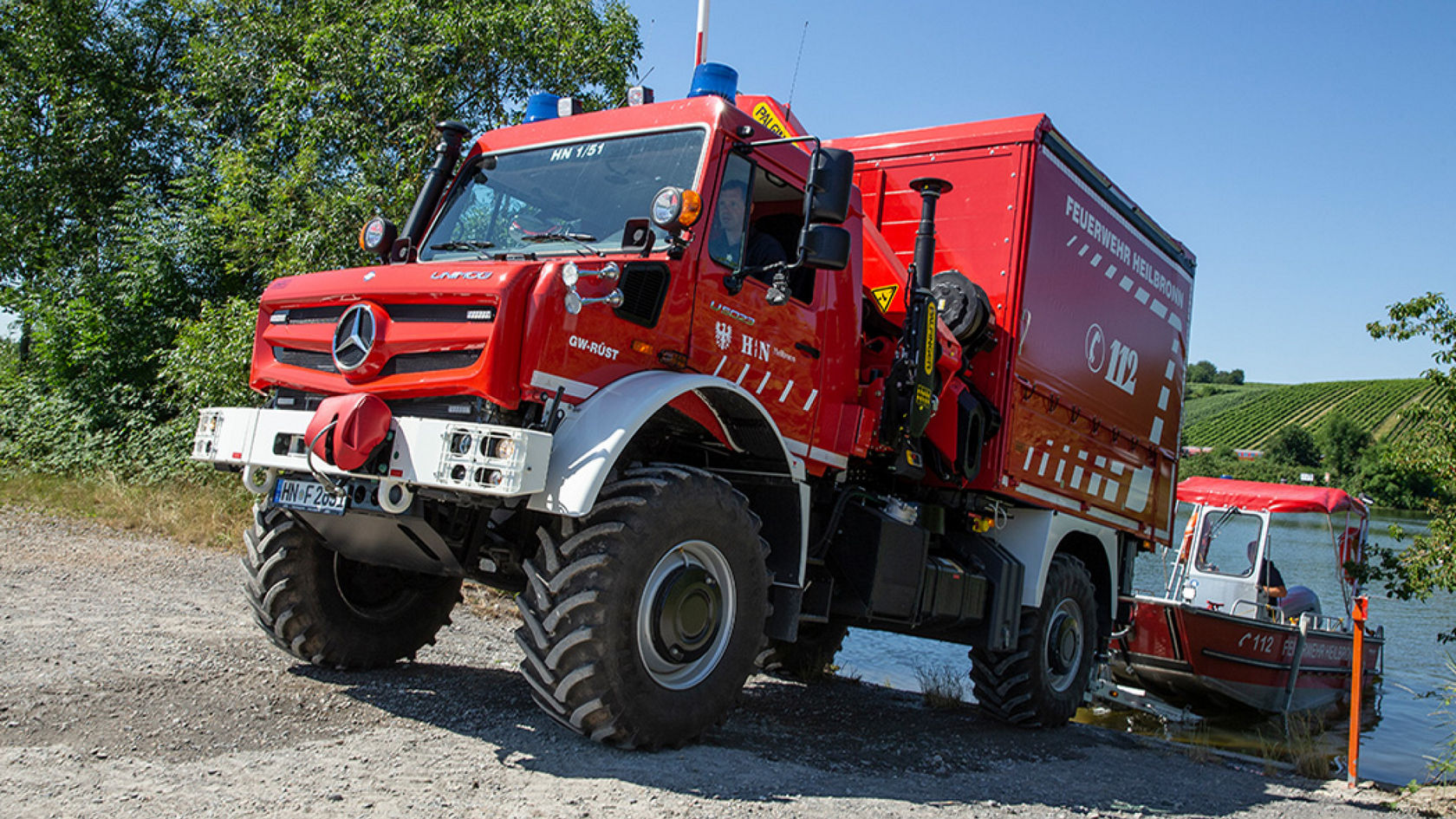 A red Unimog standing on the shore of a body of water that has launched a small, red fire-fighting vessel. The Unimog has a traditional fire service body and seems to be equipped for water rescue operations. It is standing on a gravel path. In the background you can see vines, water and trees. 