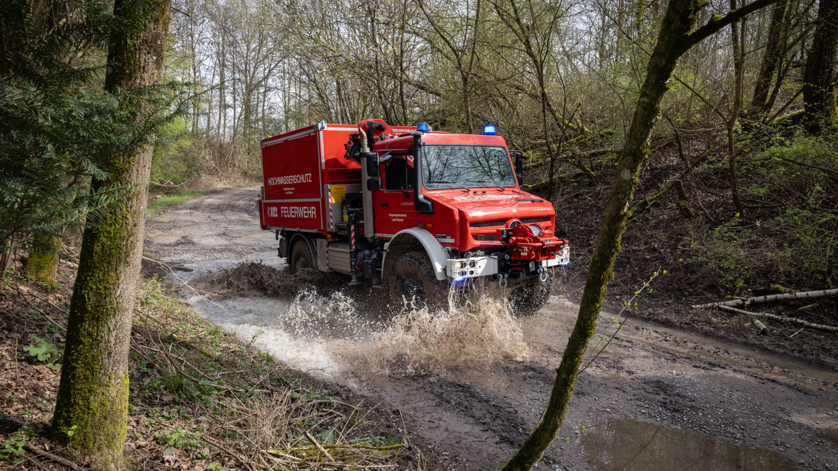  A red fire service Unimog driving on a muddy forest road. The flying spray indicates the high speed and off-road capability of the vehicle. The body of the vehicle is designed for forest fire fighting.