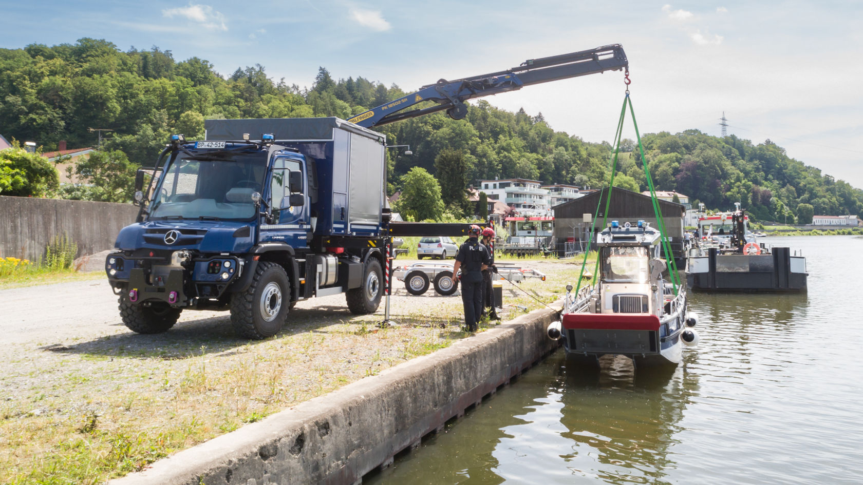 A dark blue Unimog with a crane body lifting a vessel out of the water. Two people in uniform are standing on the shore, securing the manoeuvre. A river and wooded hills can be seen in the background.