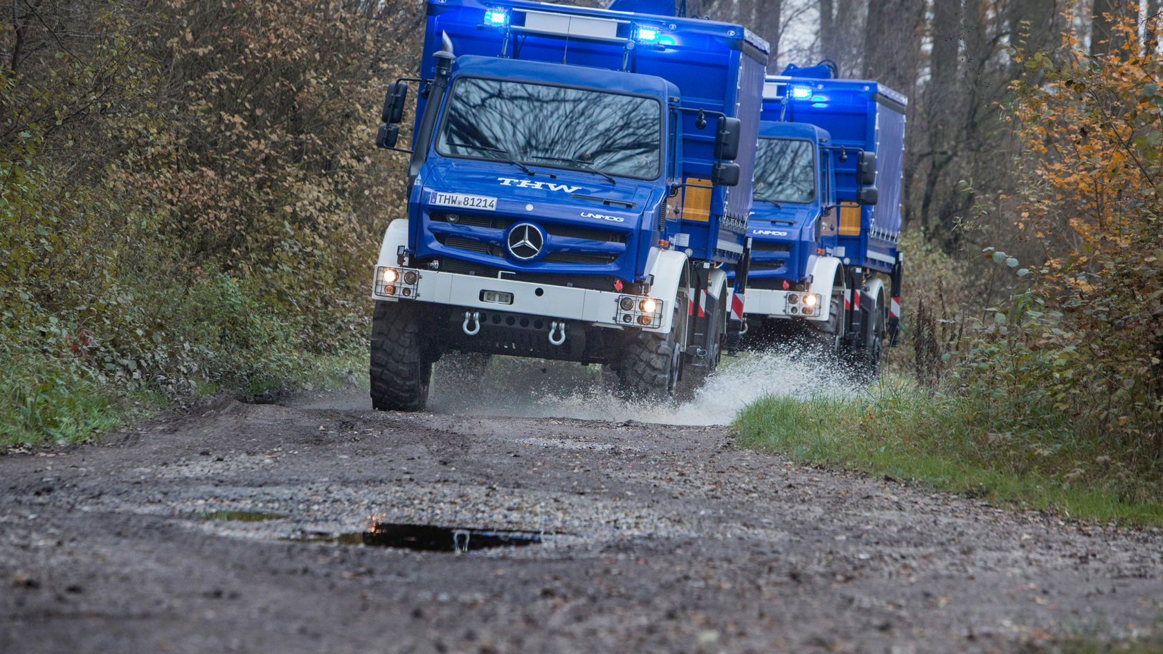 Two blue Unimogs with “THW” lettering driving with their blue lights on along a muddy forest road. They are splashing up water. 