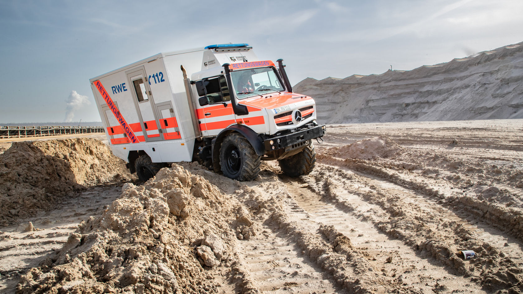 A white-and-orange rescue vehicle with “RWE” and “112” written on it making its way across a sandy industrial site. The Unimog has large tyres and high ground clearance. 