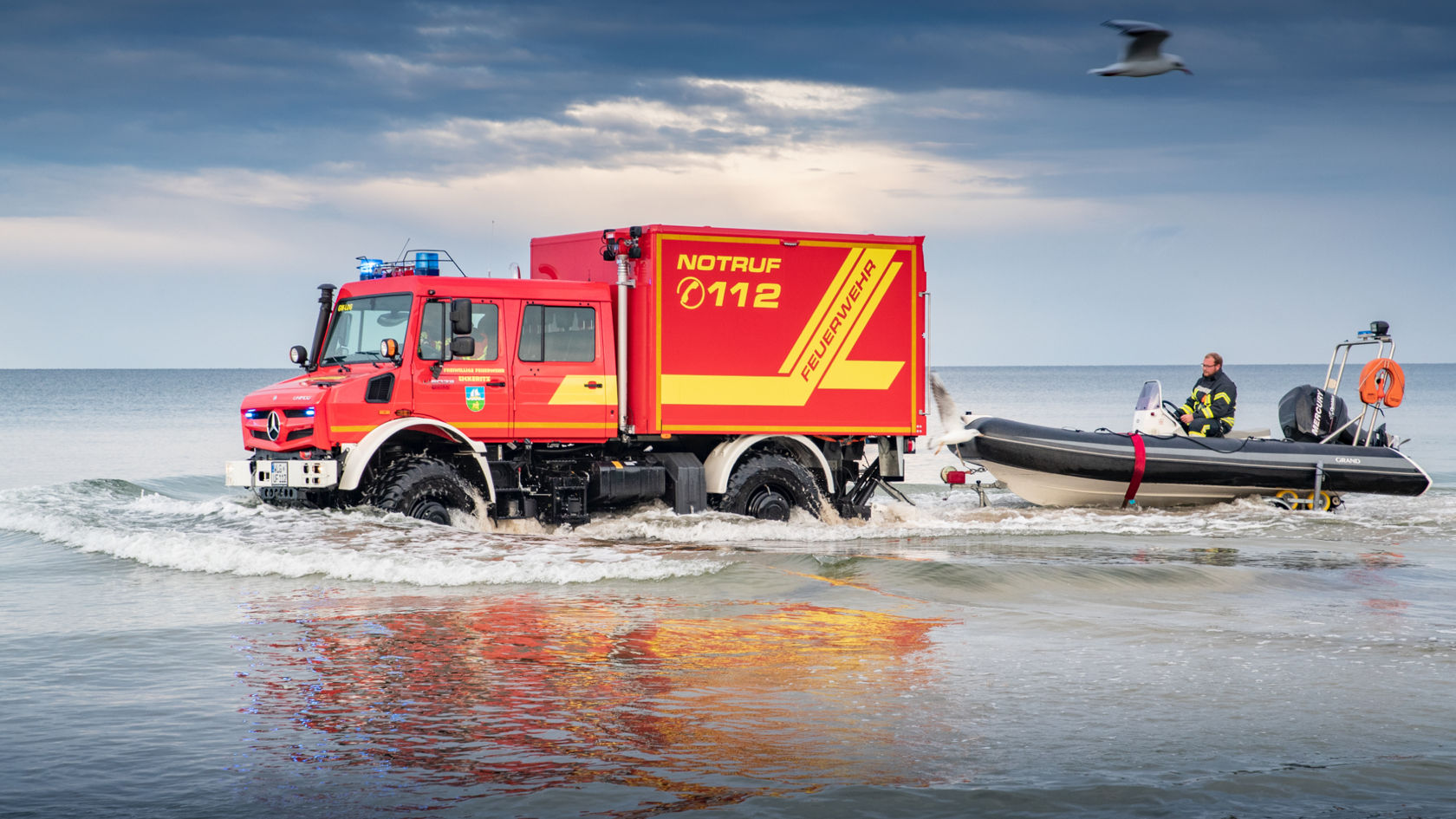 A red Unimog fire-fighting vehicle driving through shallow waters on the beach towing a rubber dinghy. A firefighter controlling the boat, while the vehicle has its blue lights on and features “NOTRUF 112” lettering. The sky is blue and there is an out-of-focus seagull in the foreground. 