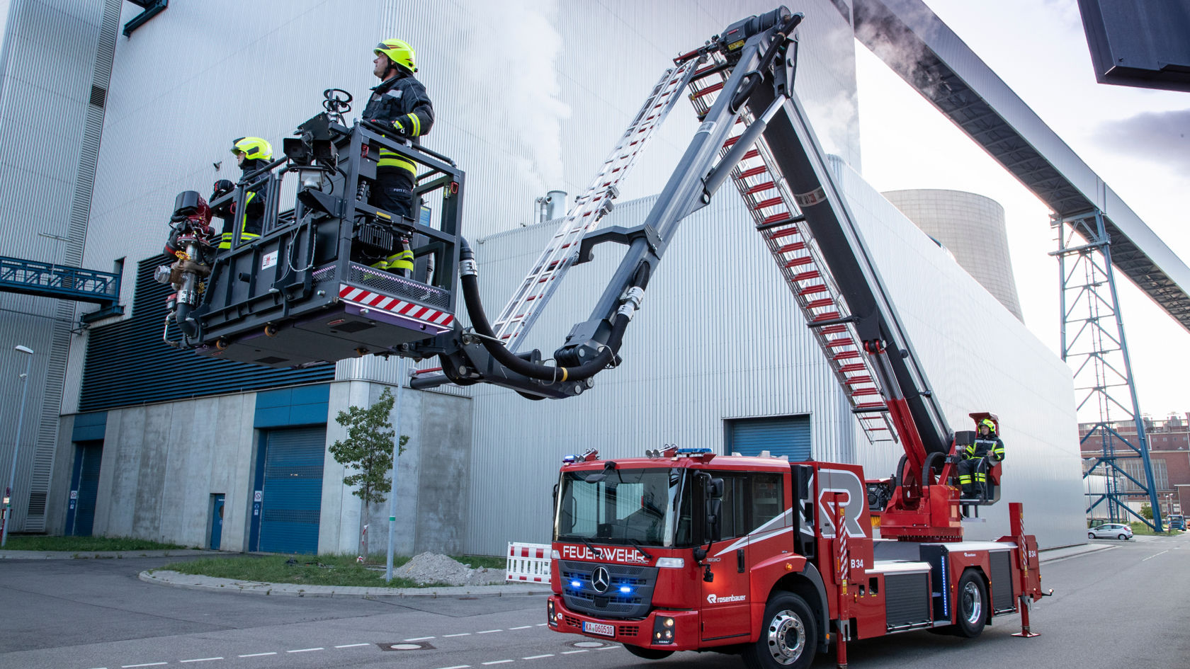 A red Econic with an extended articulated mast in front of an industrial building. Two firefighters in the cage of the aerial rescue platform operated by another crew member. The scene takes place in front of an industrial building. 