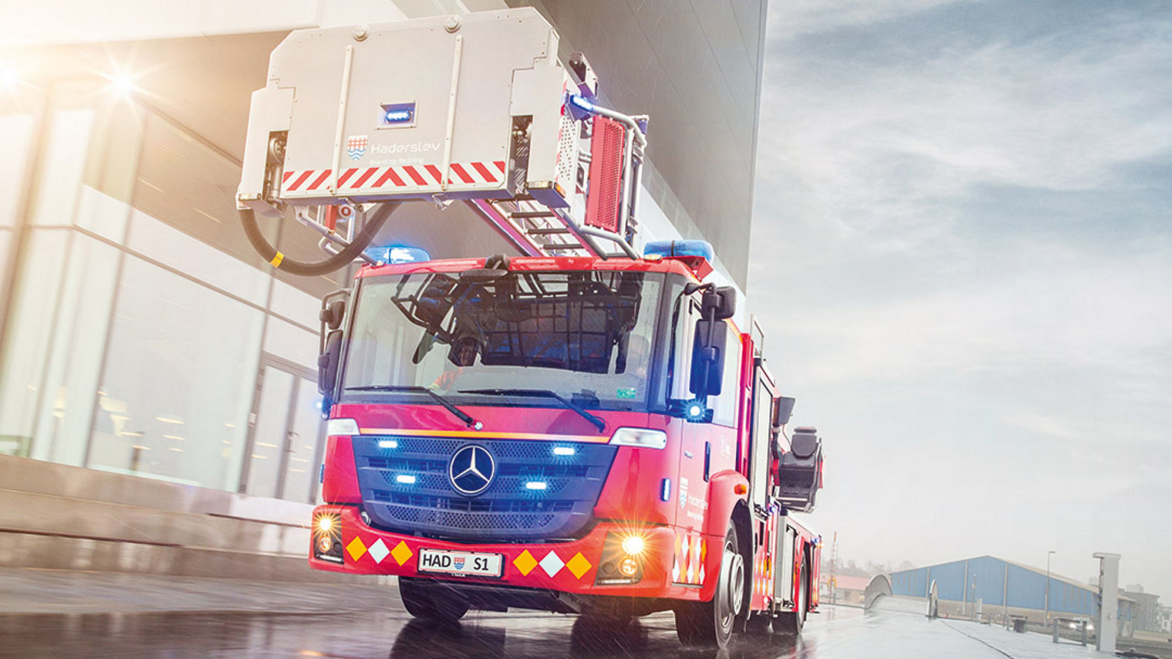 A red Econic with an aerial rescue platform driving to an incident on a rainy road, surrounded by industrial buildings. The blue lights of the fire-fighting vehicle is switched on.